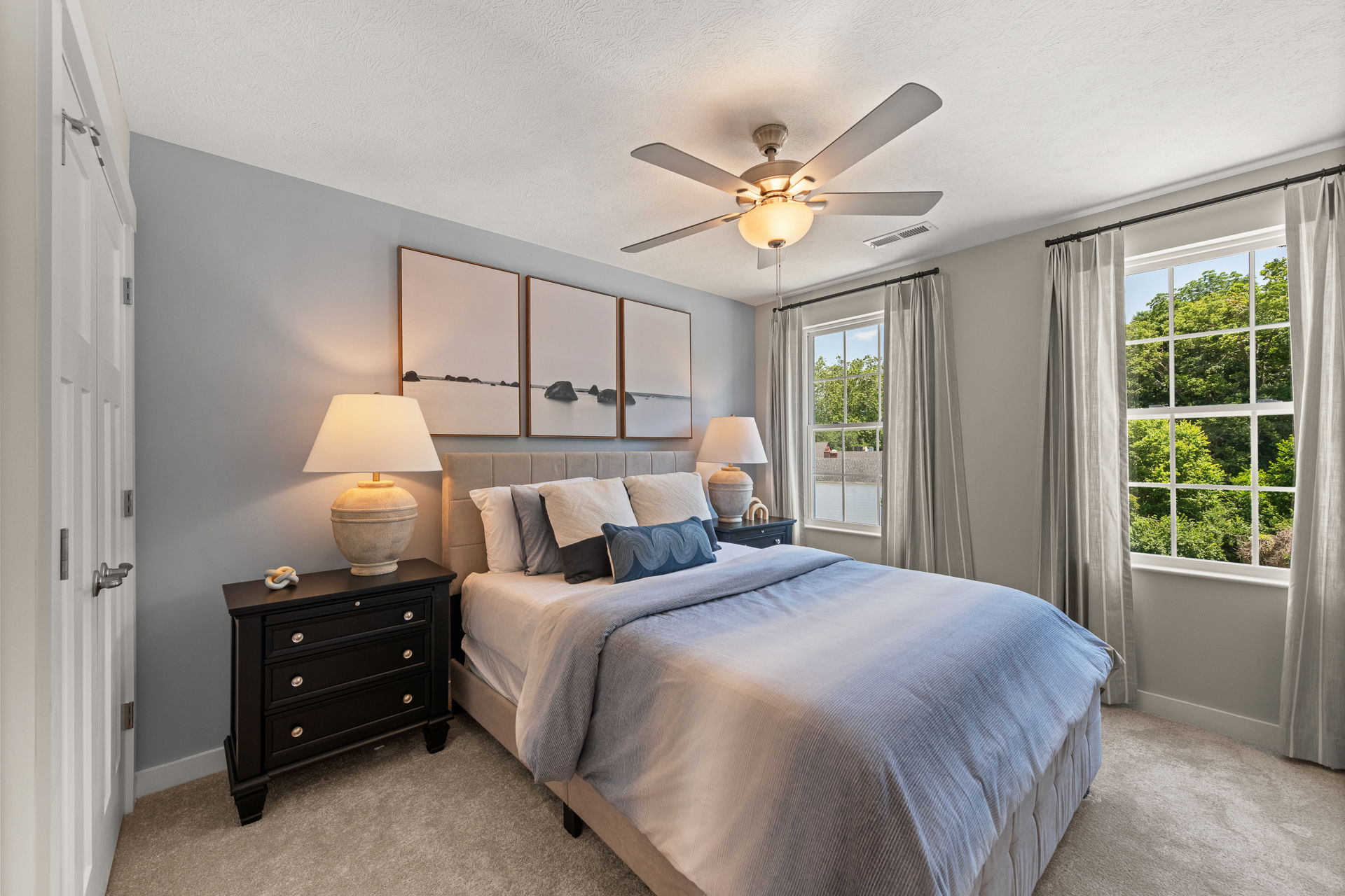 Modern bedroom with blue and white decor, featuring a cozy bed, elegant nightstands with lamps, and large windows overlooking greenery.
