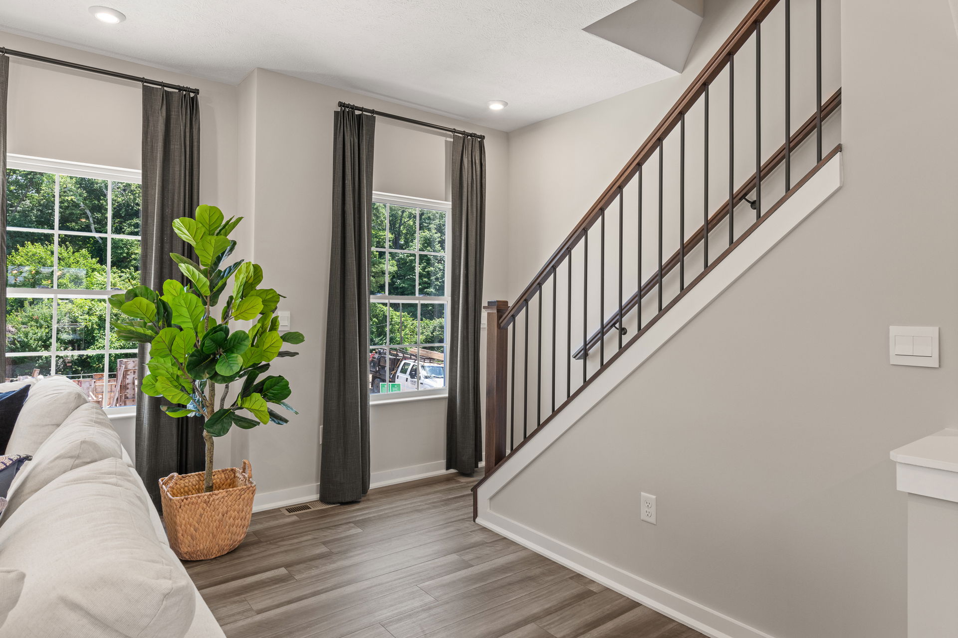 Modern living room interior with a gray sofa, large windows with dark curtains, a staircase with a metal railing, and a potted plant in a woven basket.