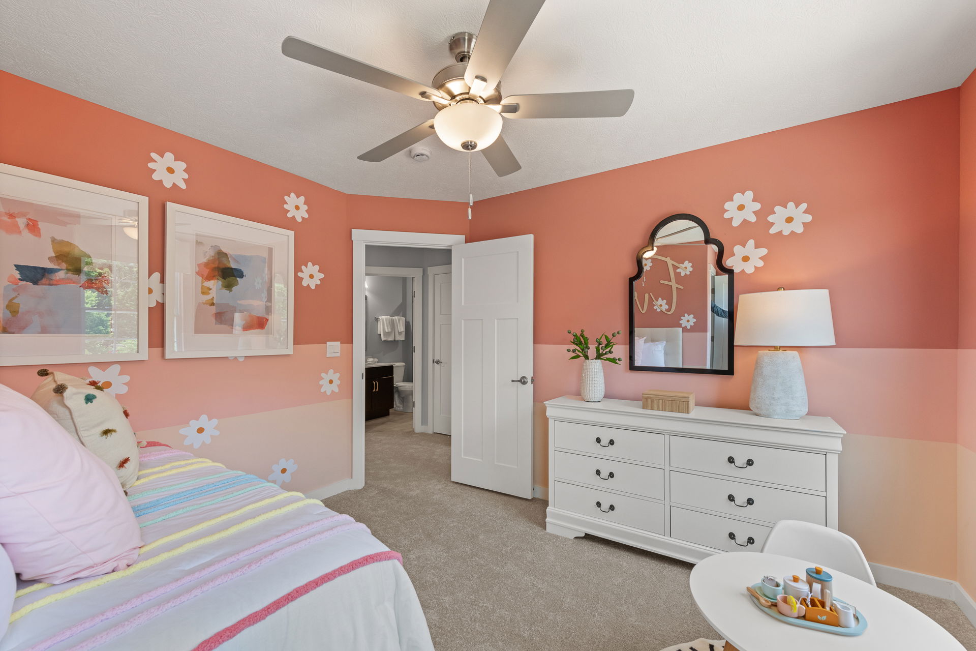 Brightly decorated children's bedroom with coral walls adorned with white daisy decals, a ceiling fan, white furniture, and colorful bedding.