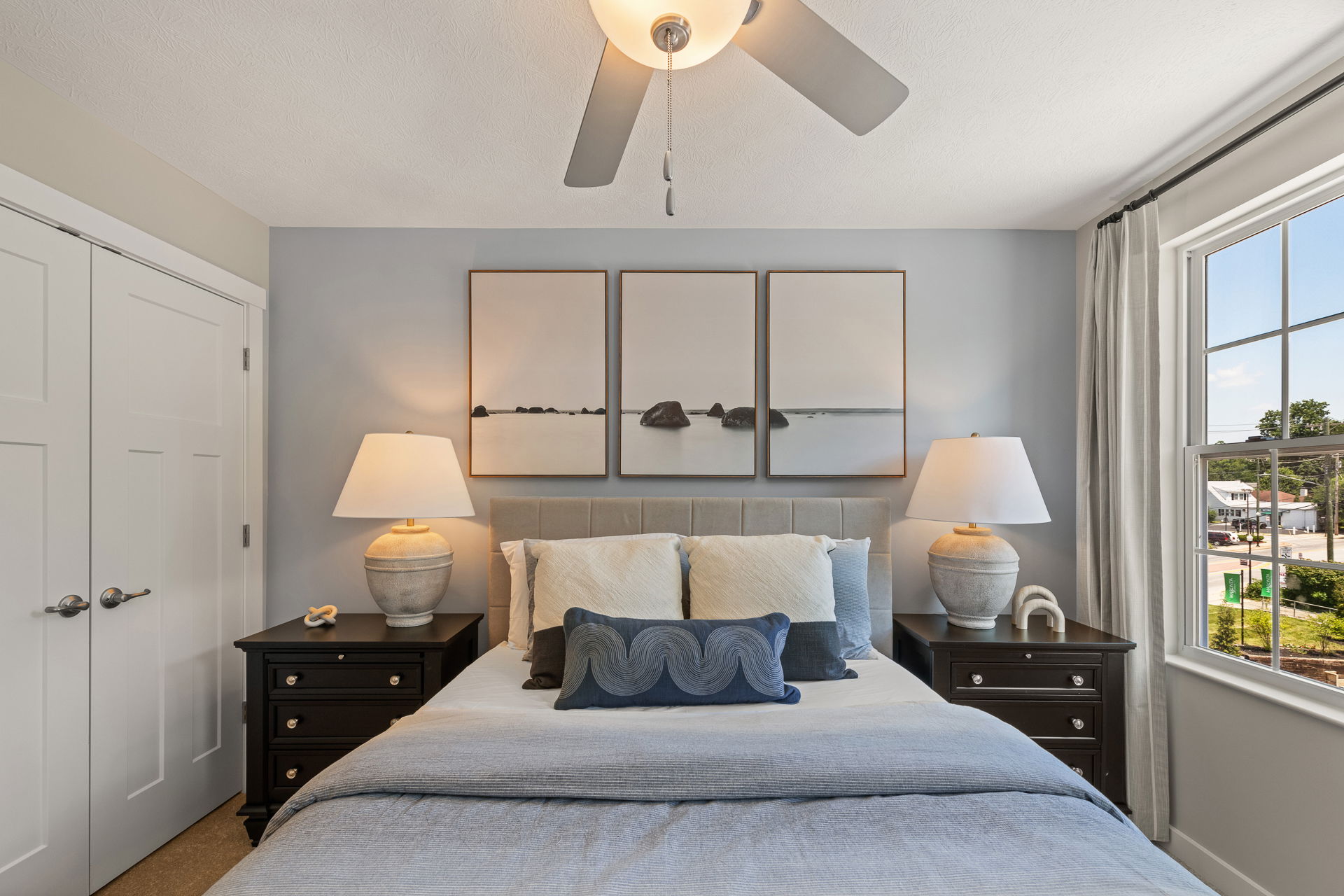 Modern bedroom with a neatly made bed, decorative pillows, matching side lamps, and a serene triptych wall art above the headboard.