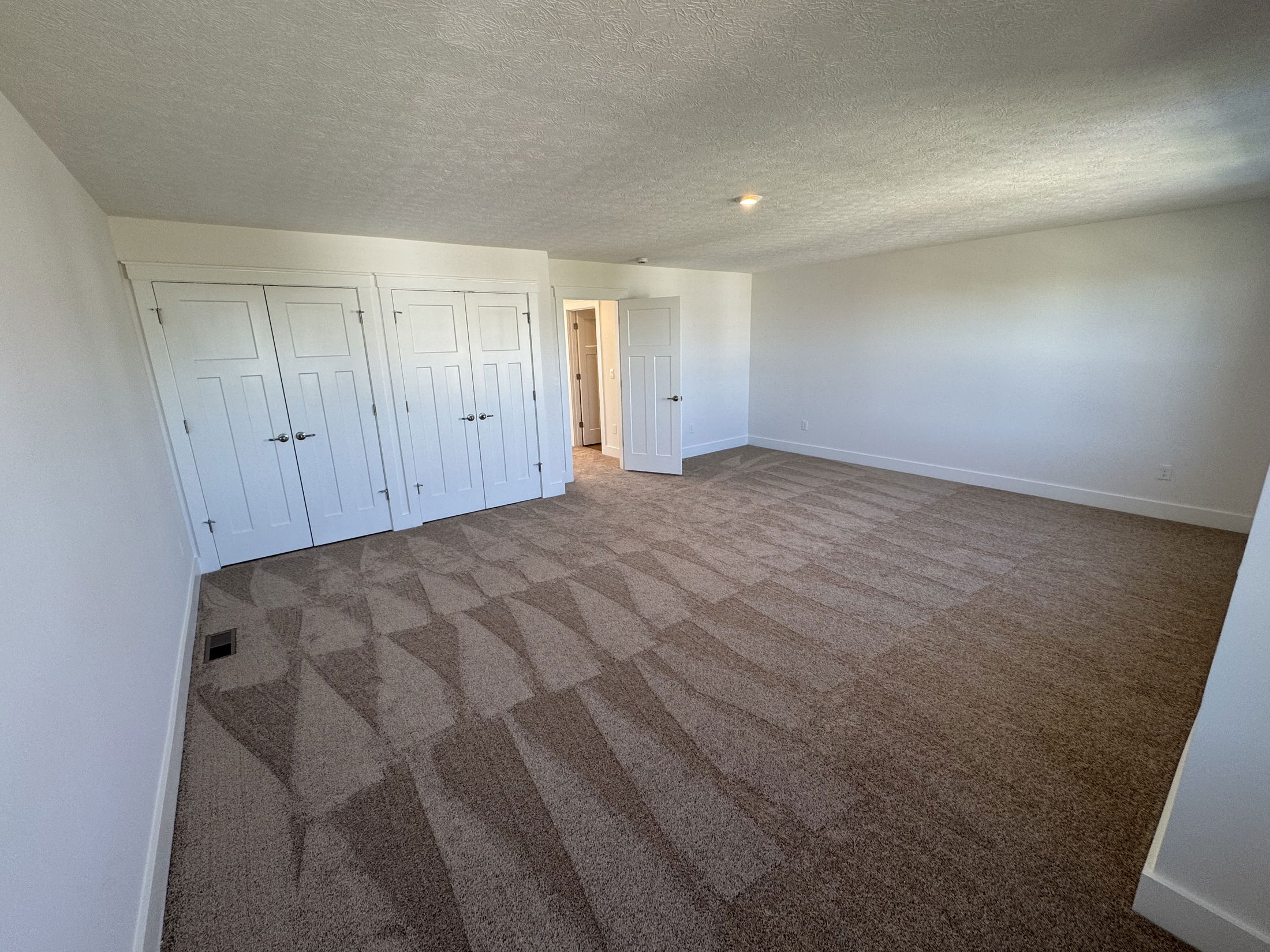 Spacious and bright empty bedroom with new carpet, double white sliding doors, and natural light coming from nearby doorways.