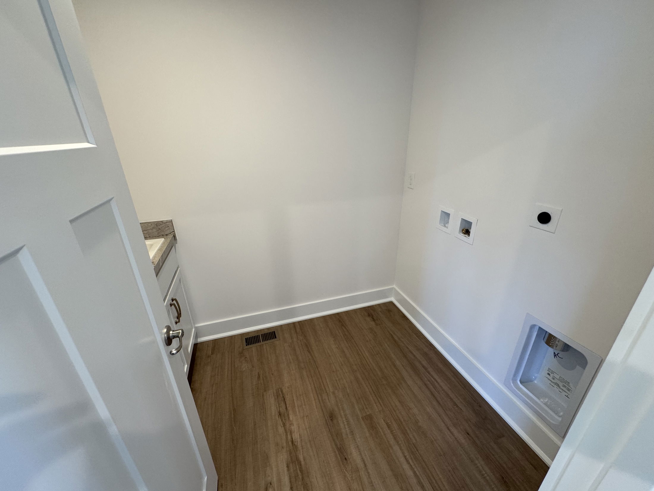 Modern laundry room with white cabinetry and hardwood flooring, featuring electrical outlets and plumbing connections.
