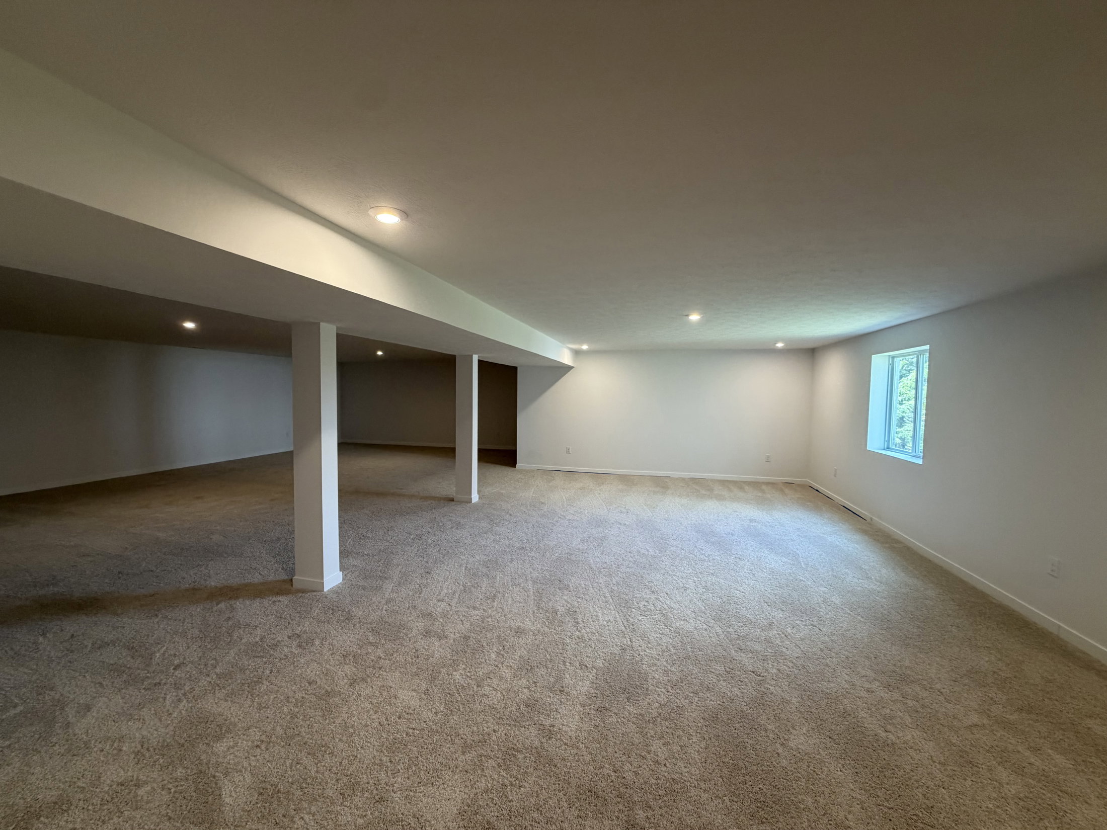 Spacious, empty basement with light-colored walls, carpeted floor, and a window letting in natural light.