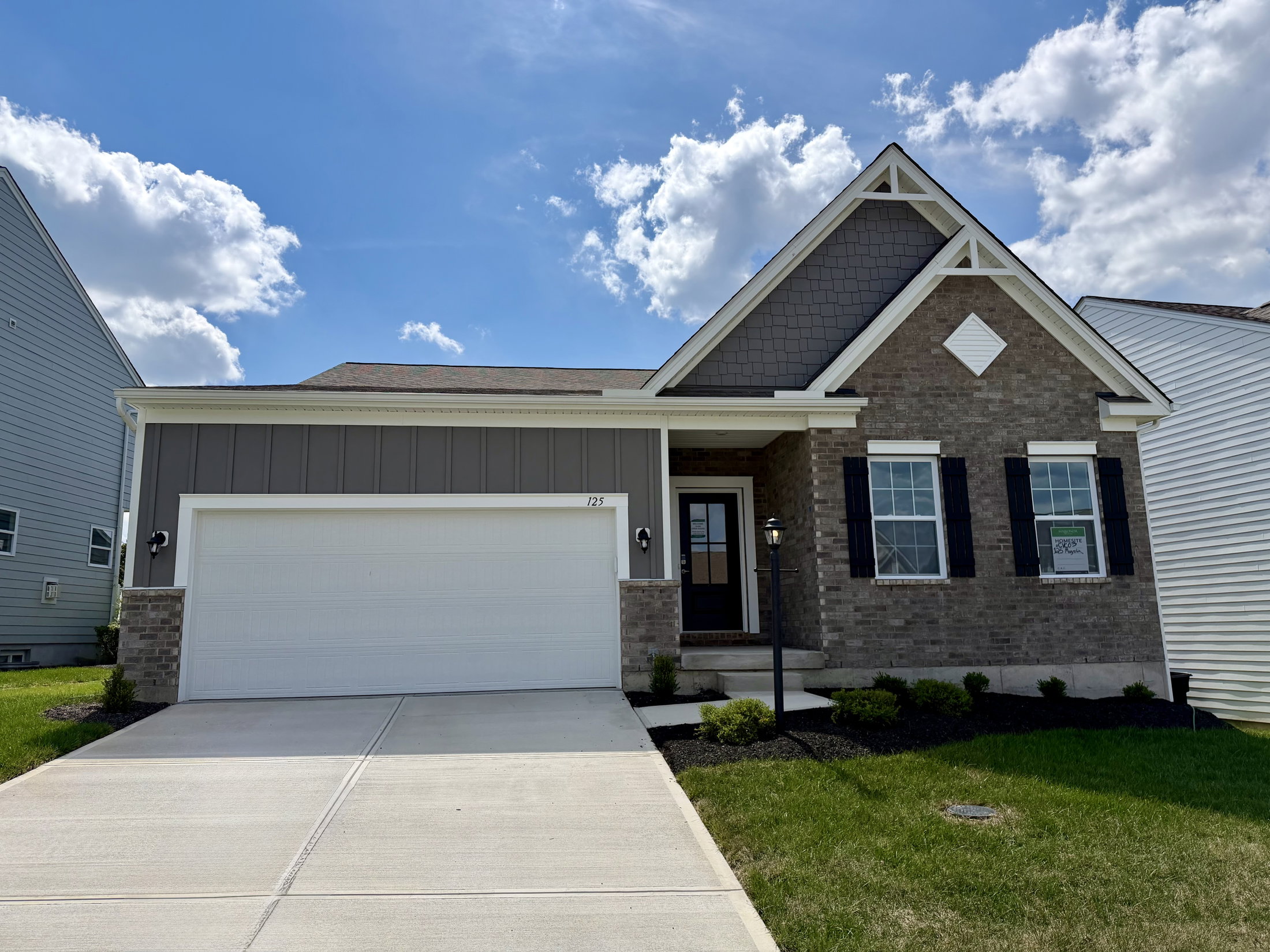 A single-story house with a gray and brick exterior, black shutters, and a two-car garage, surrounded by a lawn under a partly cloudy sky.