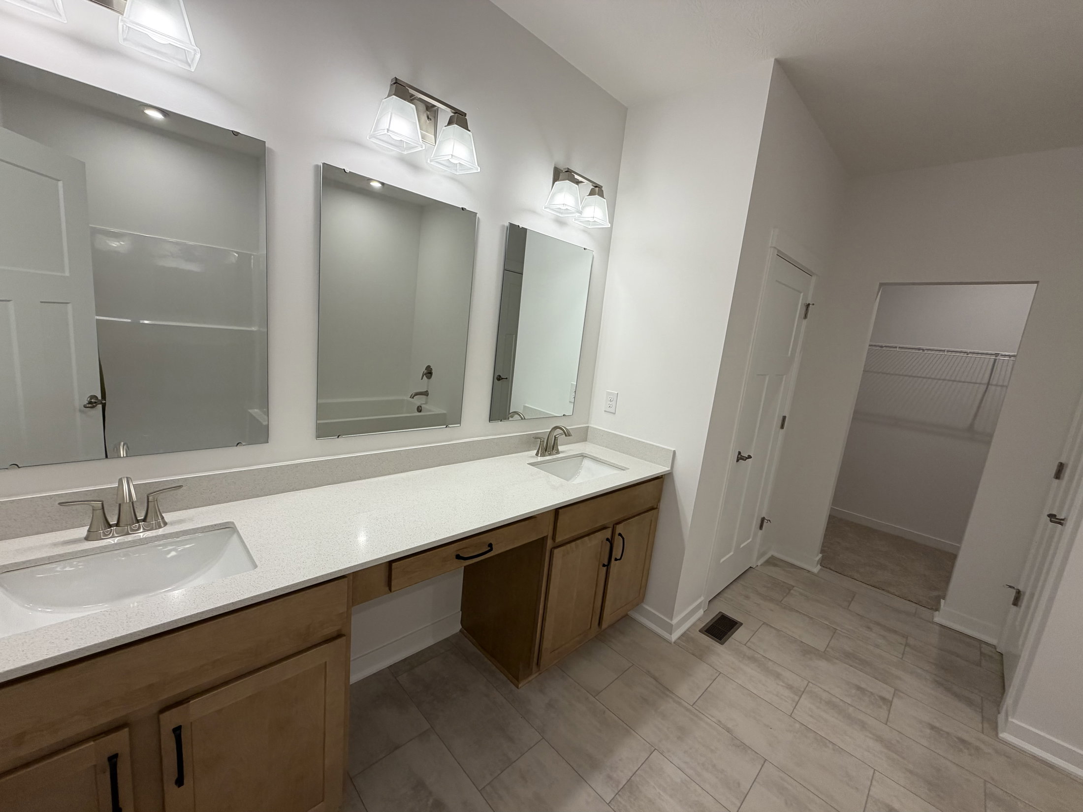 A well-lit bathroom featuring two sinks, three mirrors, and a doorway leading to a closet.