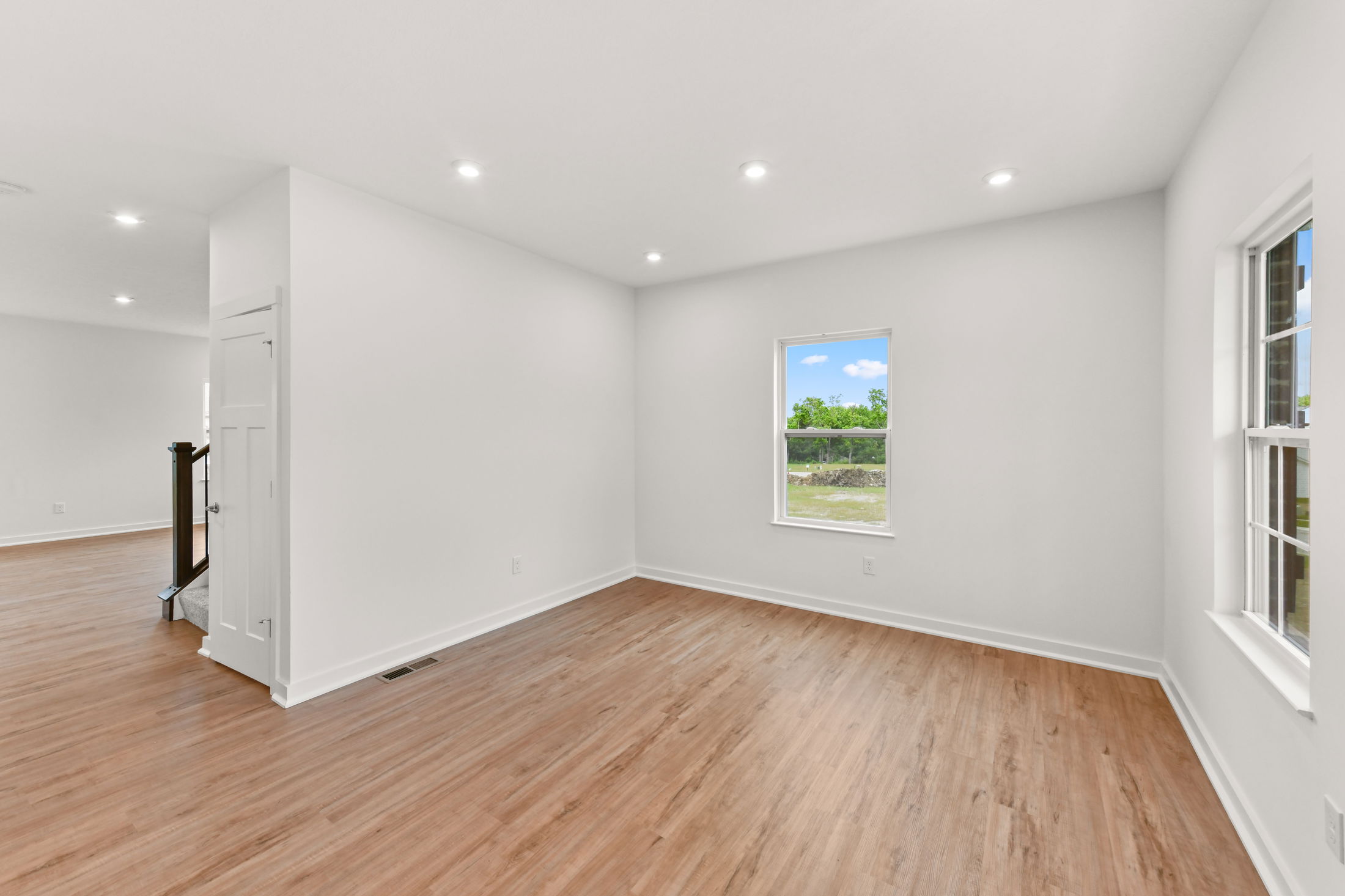 A bright, empty room with white walls, hardwood floors, recessed lighting, and large windows offering a view of the greenery outside.