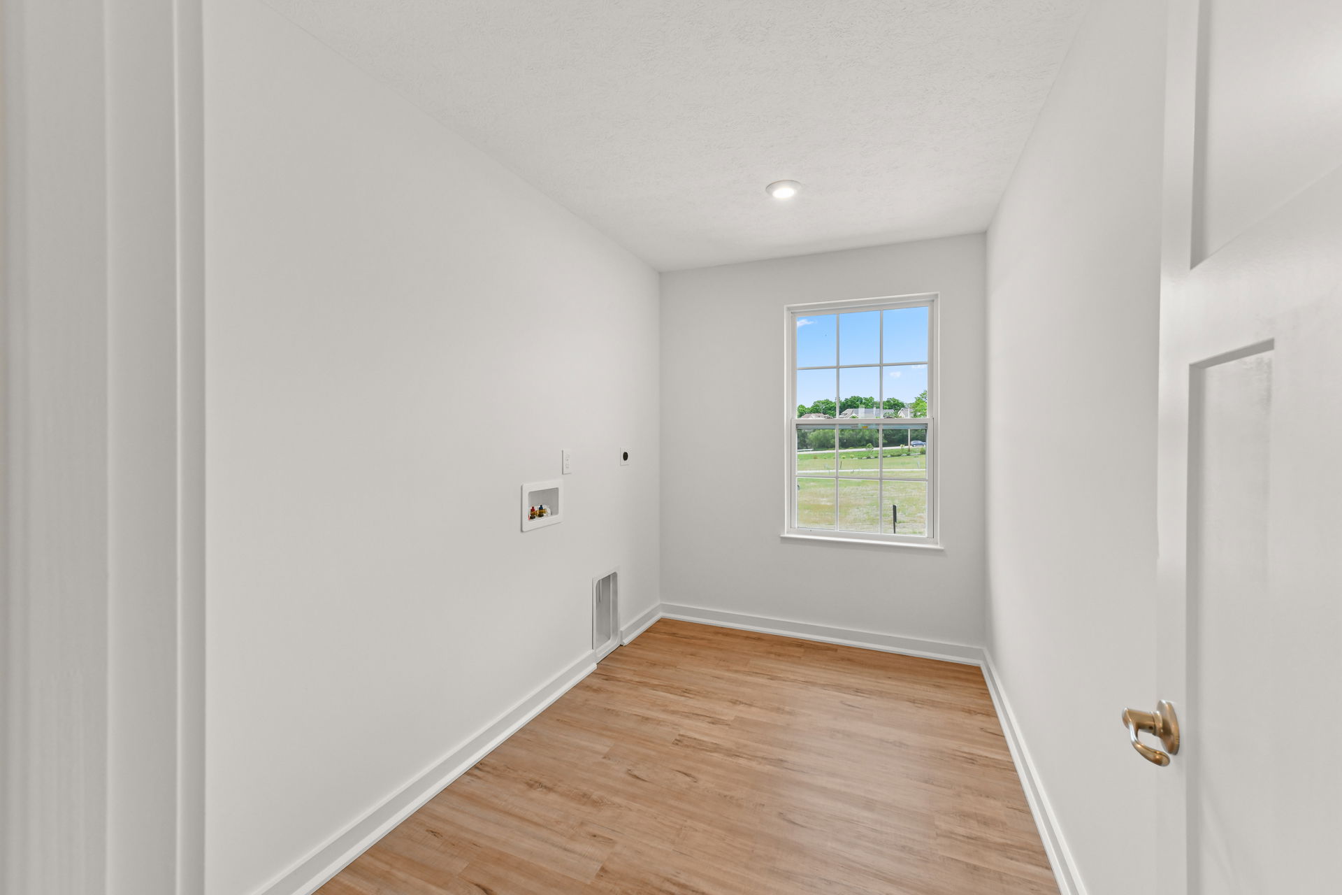 Bright, empty laundry room with white walls, wood flooring, and a large window overlooking a grassy landscape.