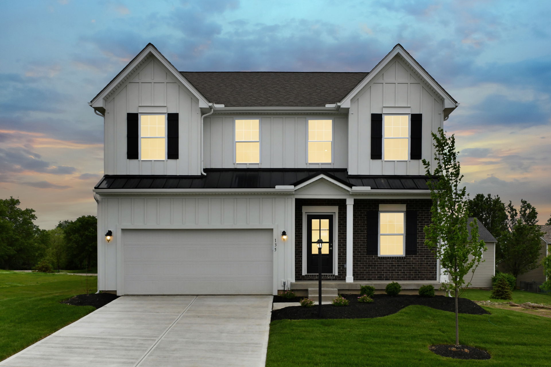 Modern two-story house with a well-lit facade, dark shutters, garage, and landscaped front yard at dusk.