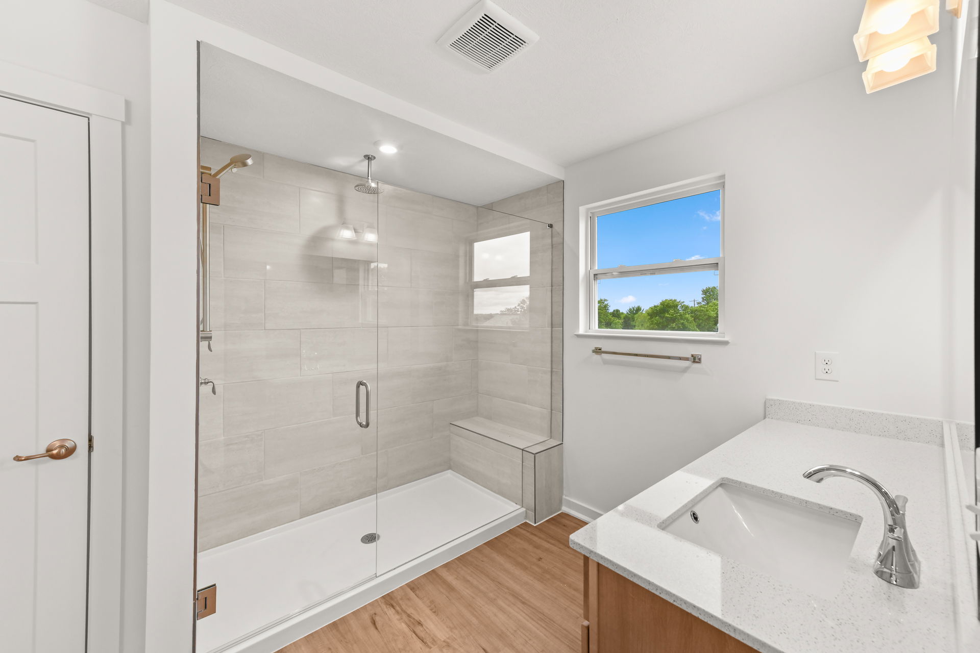 Modern bathroom with a spacious glass-enclosed shower, large window, and sleek white countertop sink.