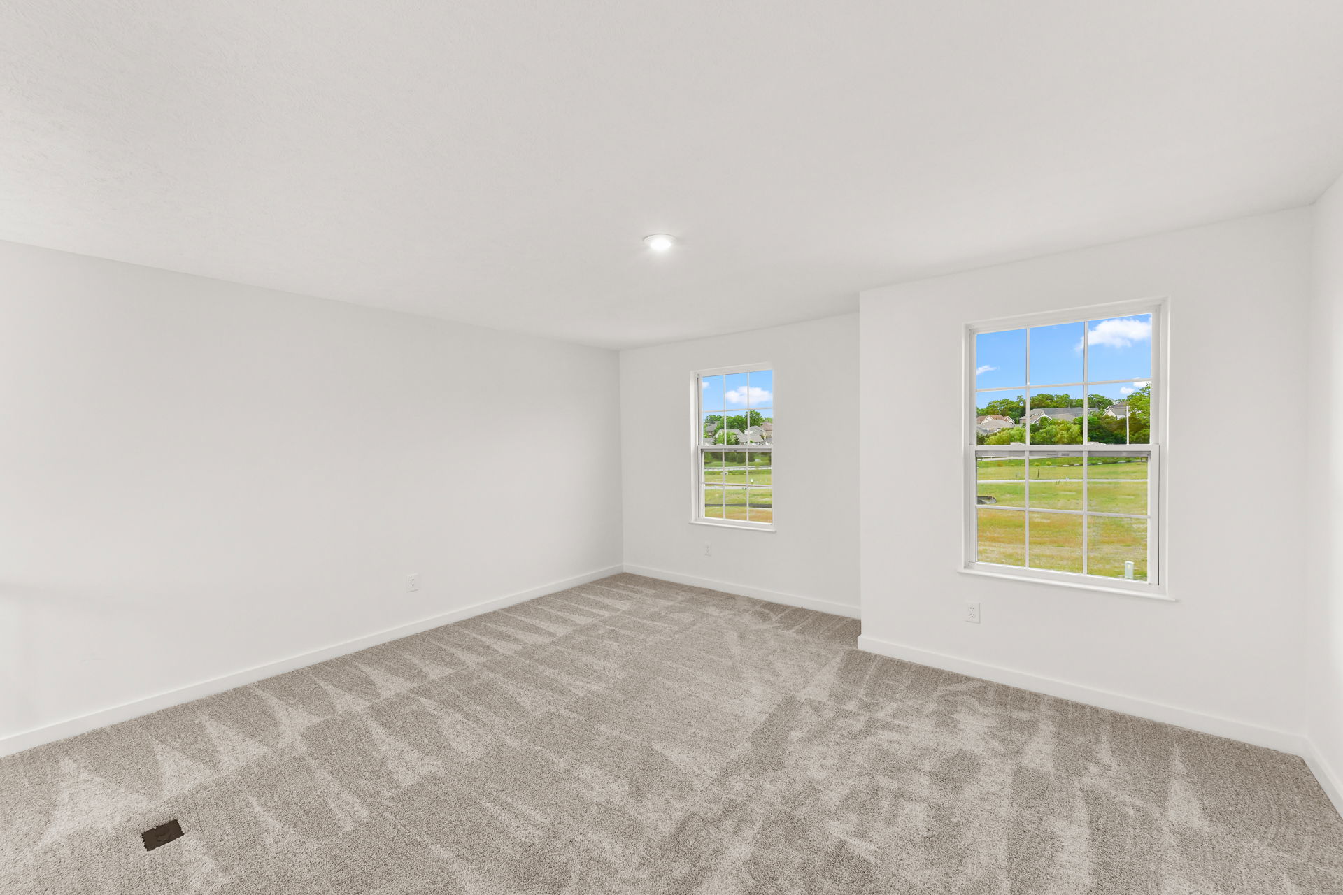 A spacious, empty room with white walls, gray carpet, and two large windows overlooking a grassy landscape.