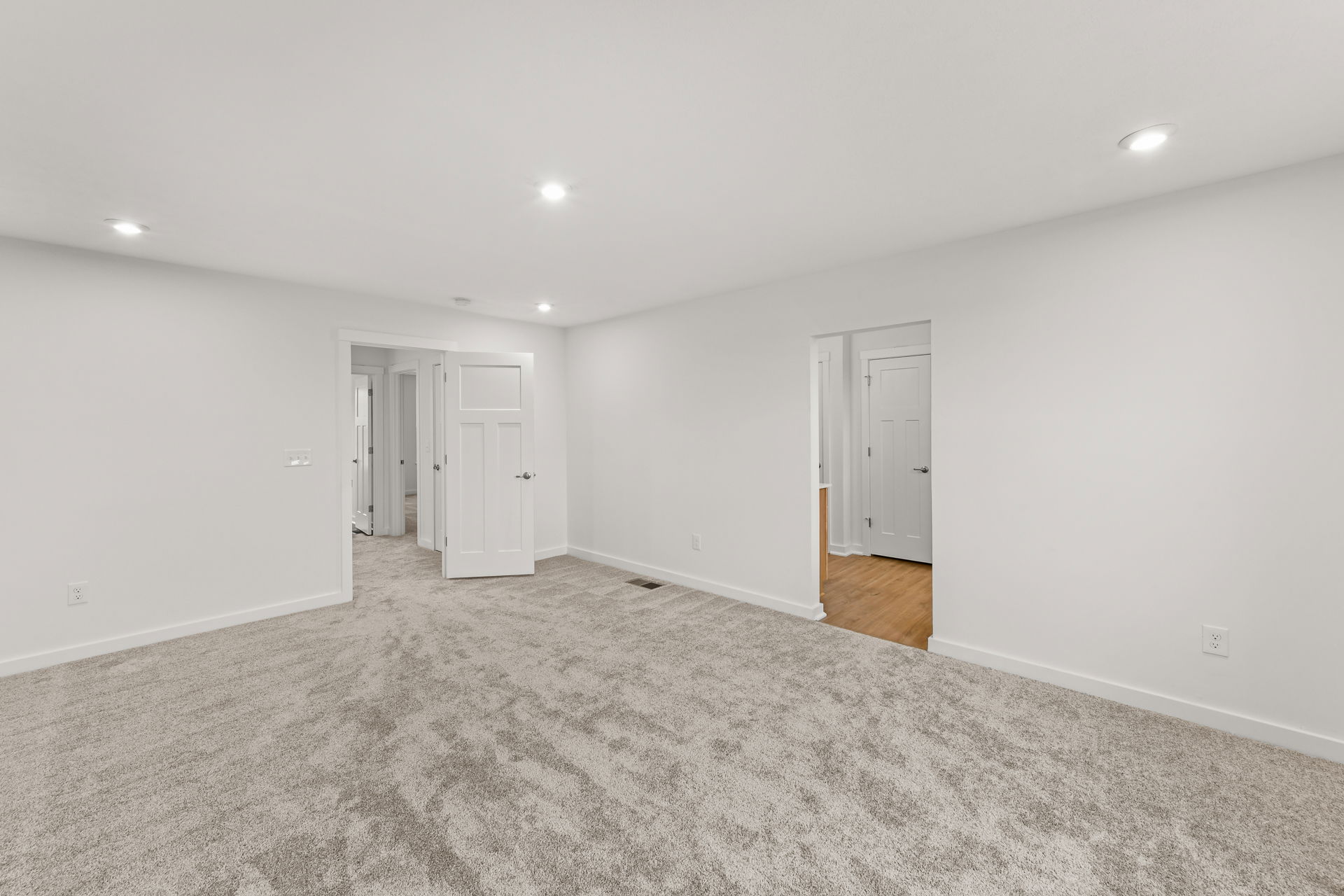 Spacious empty room with clean white walls, recessed lighting, and freshly installed gray carpeting.