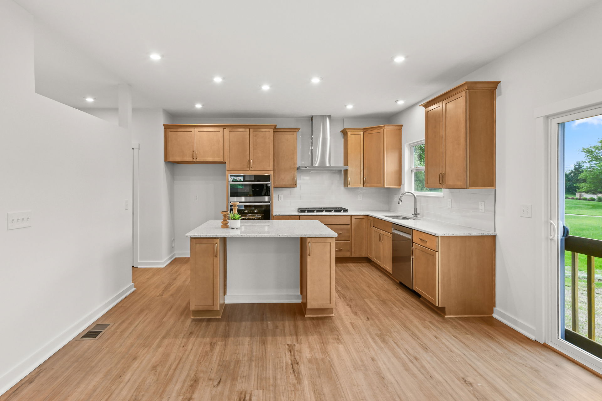 Modern kitchen with light wood cabinets, center island, stainless steel appliances, and a view of greenery through the window.