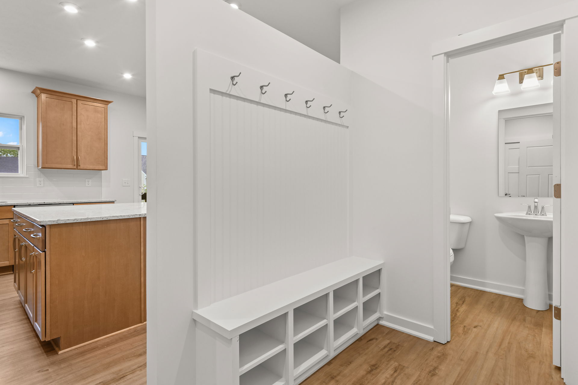 Modern mudroom with white storage bench, coat hooks, and adjacent bathroom, featuring wooden flooring and a view of the kitchen with light wood cabinetry and a granite countertop.