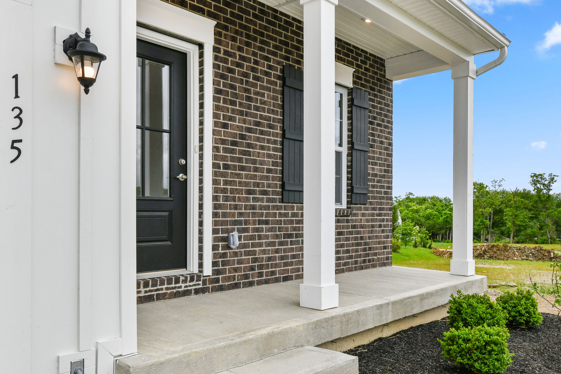 Front porch of a modern brick house with black door and white columns, set against a green landscape.