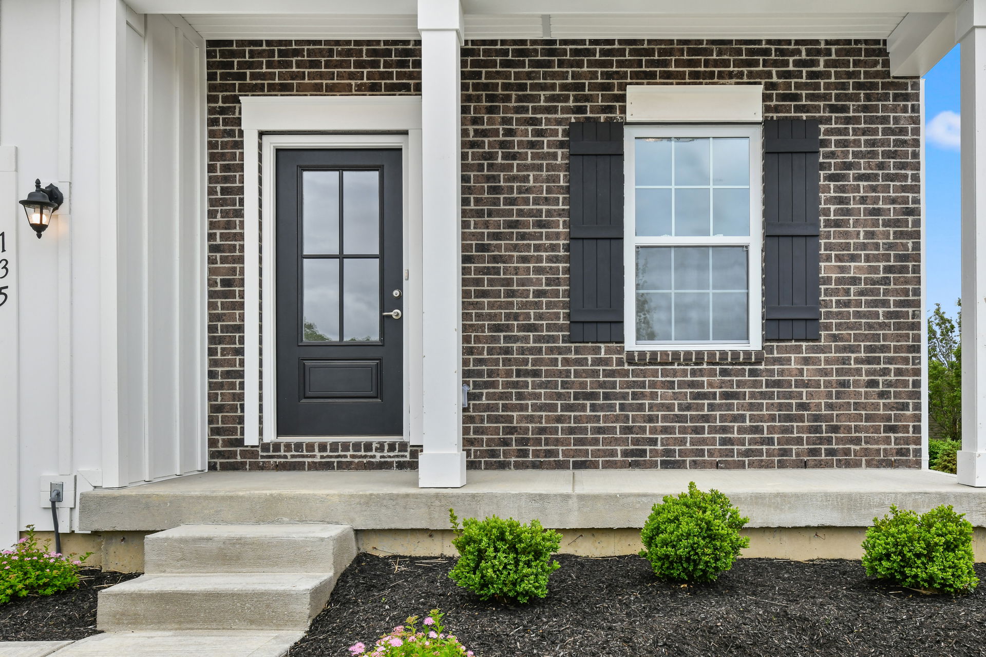 Front view of a brick house entrance with a black door, window with shutters, and manicured shrubs.