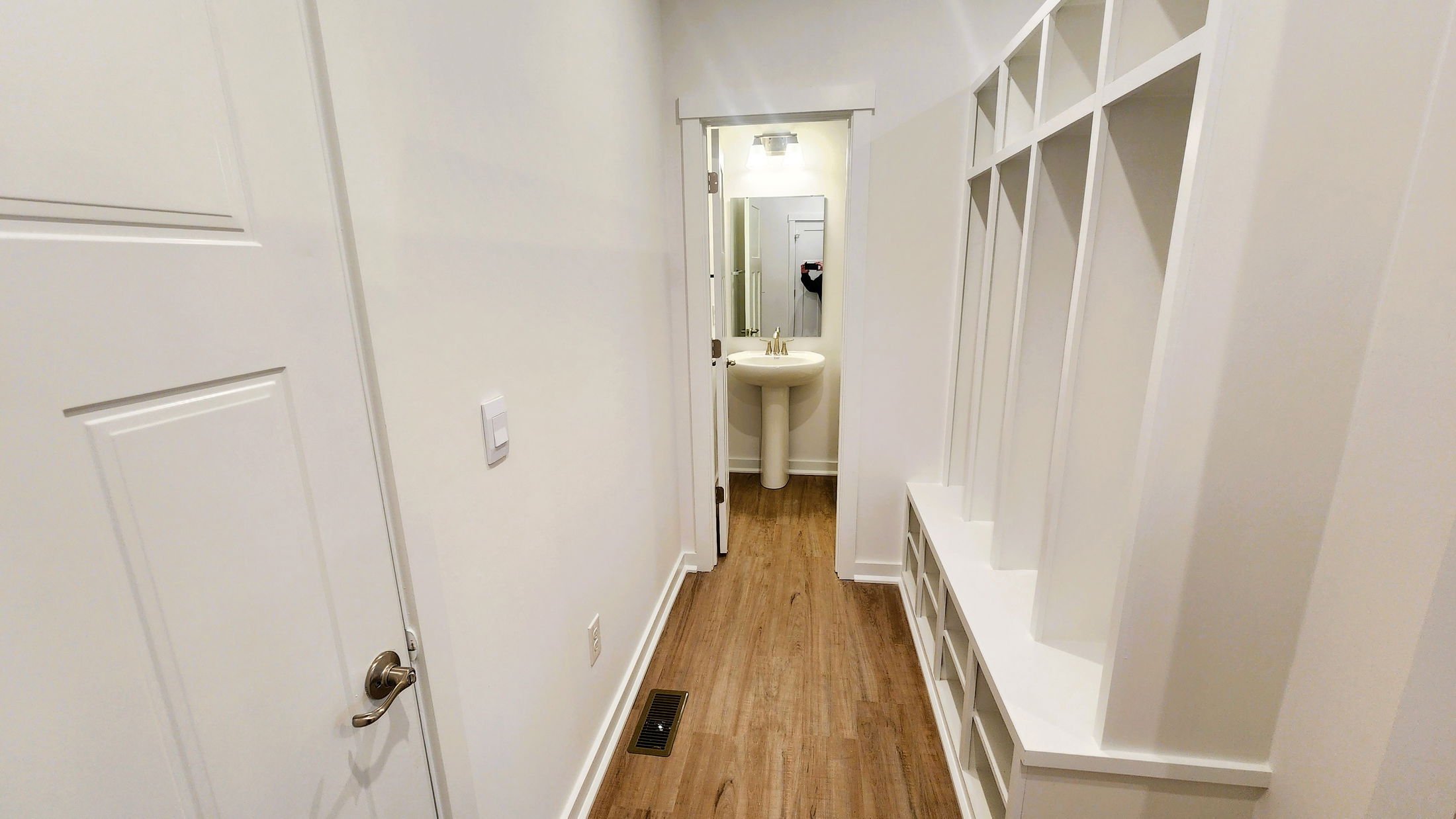 A modern hallway with a white wall, wooden flooring, and a half bathroom visible through an open door.