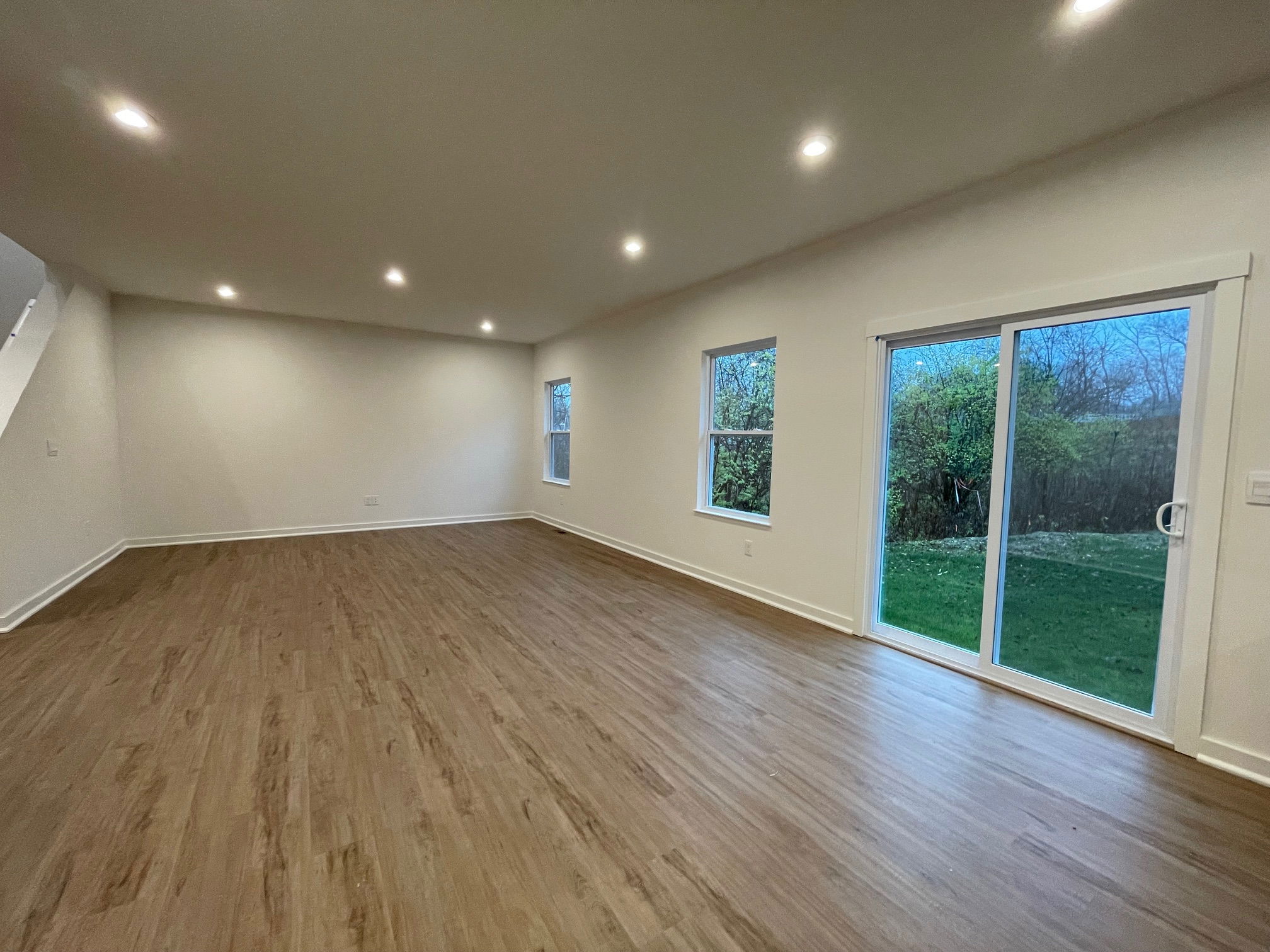Spacious empty room with hardwood flooring, recessed lighting, and a large sliding glass door overlooking a grassy outdoor area.