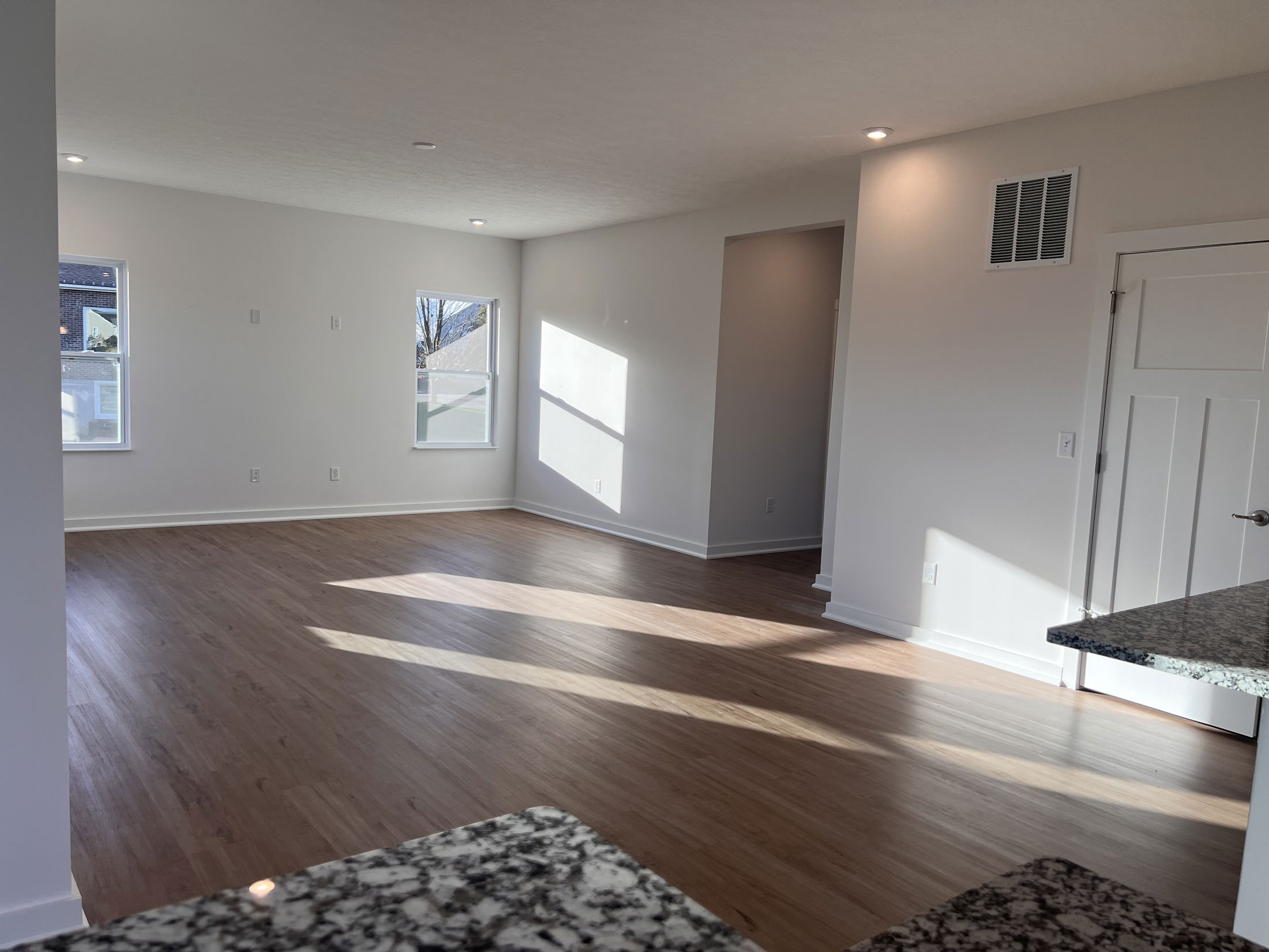 Bright, empty living room with hardwood floors, two windows, and a view of the granite kitchen counter in a modern home.