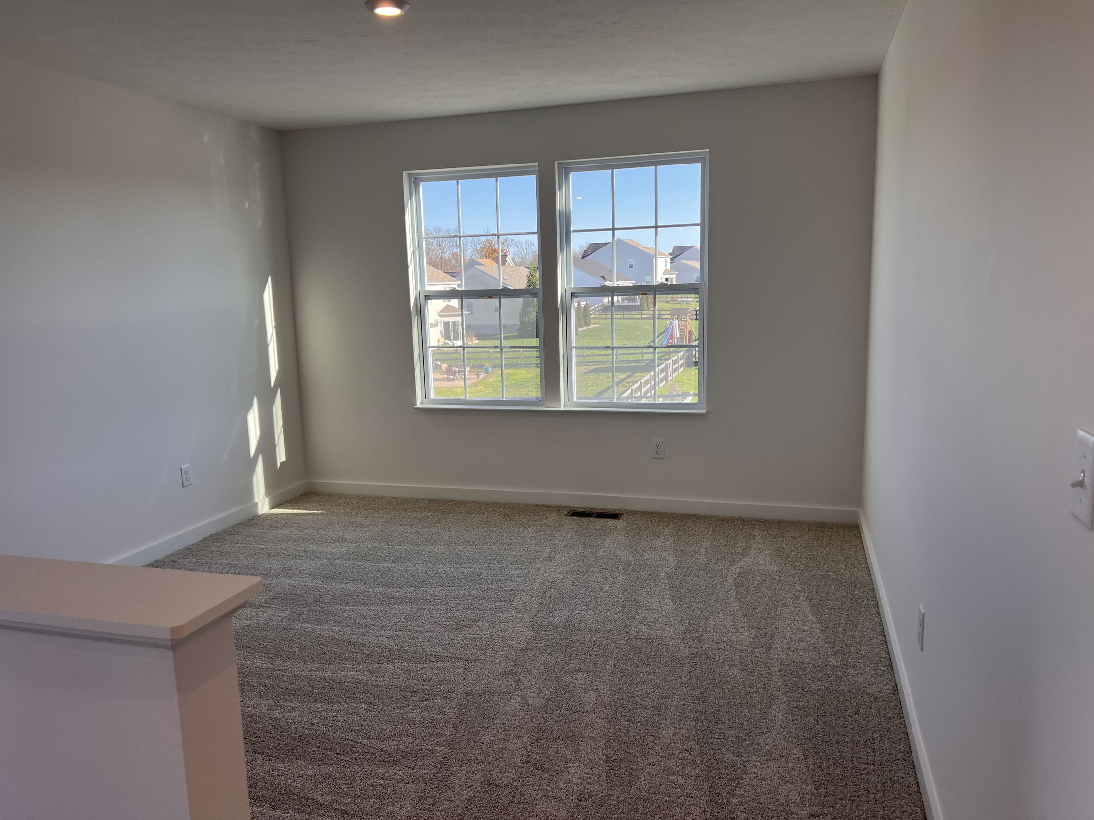 Bright, empty room with beige carpeting, white walls, and a window overlooking a suburban backyard.