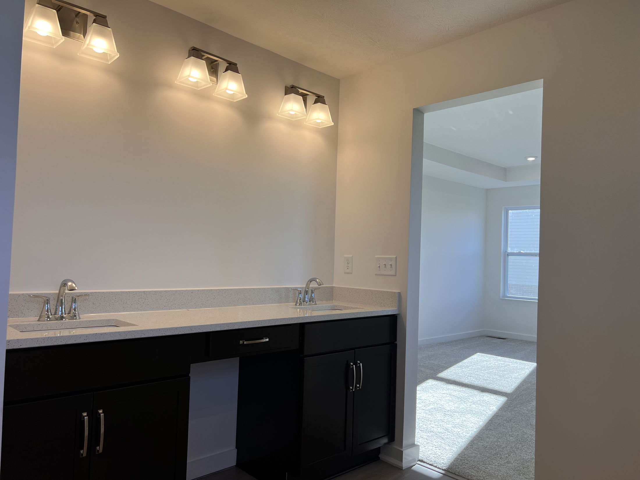 Modern bathroom with dual sinks, dark cabinetry, and bright lighting leading into a sunlit room with a large window.