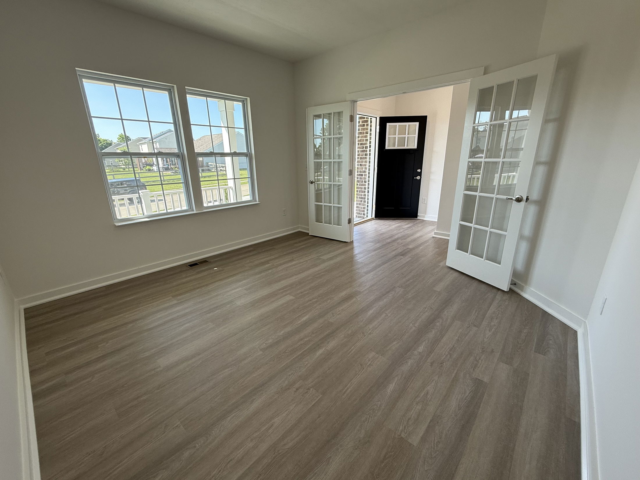 Bright, empty room with hardwood floors, white French doors, and large windows, connected to an entrance featuring a black door with glass panels.