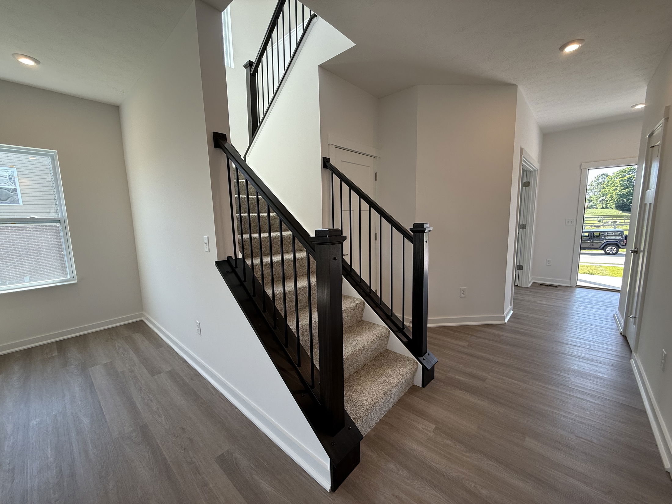 Modern home staircase with dark wood railing and carpeted steps, adjacent to a bright hallway with hardwood flooring.