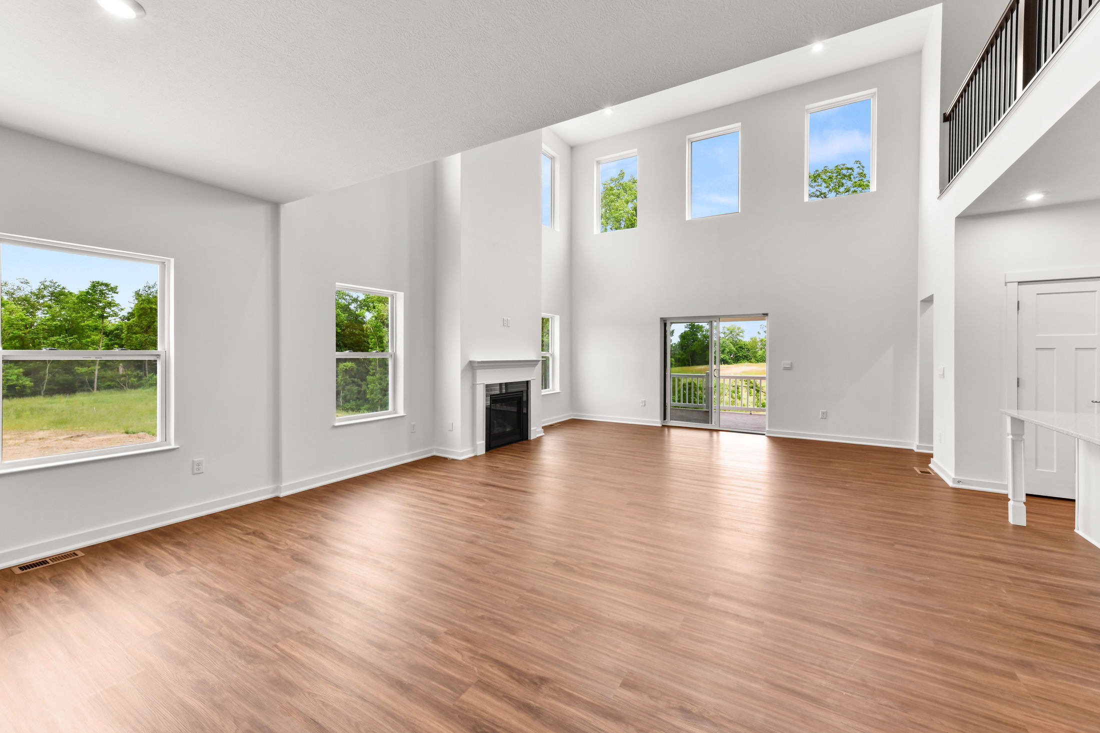 A spacious, modern living room with high ceilings, hardwood floors, large windows, and a fireplace, offering abundant natural light and outdoor views.