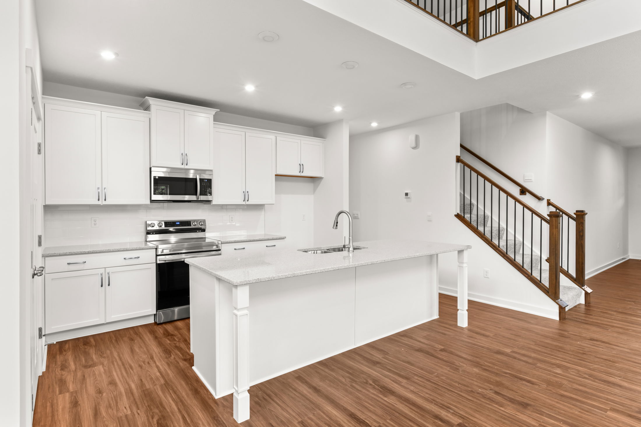 Modern kitchen interior with white cabinets, a large island, and stainless steel appliances, featuring wooden flooring and a staircase.