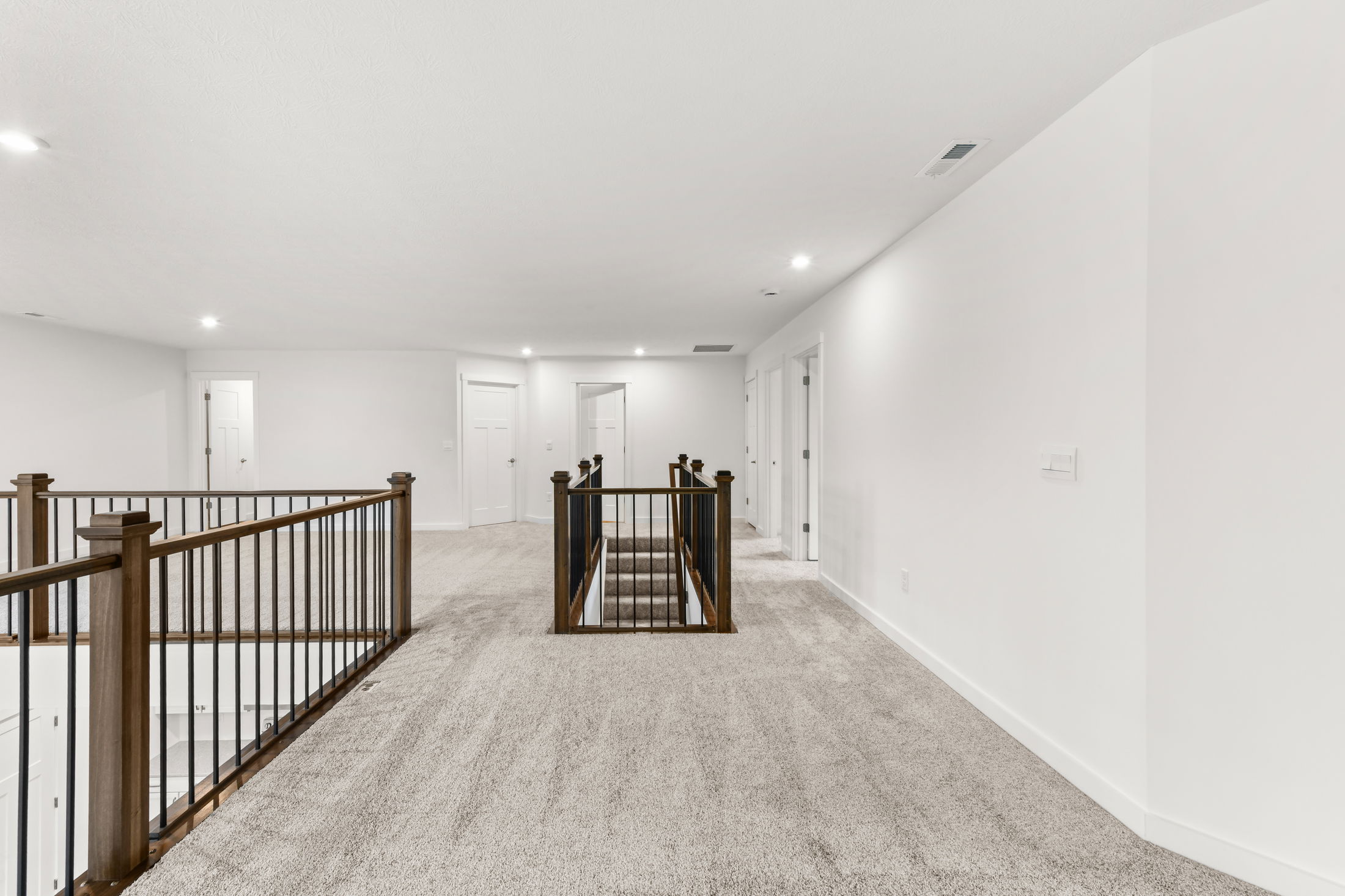 Bright, modern upstairs hallway with light gray carpet and dark wooden railing in a newly constructed home.
