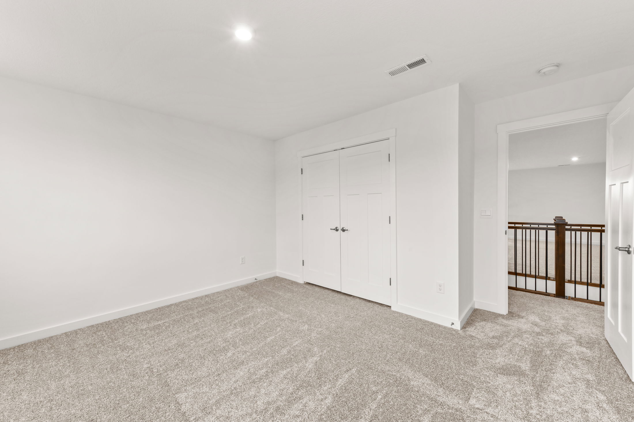 Empty room with white walls, carpeted floor, a closed closet, and an open door leading to a hallway with a wooden railing.