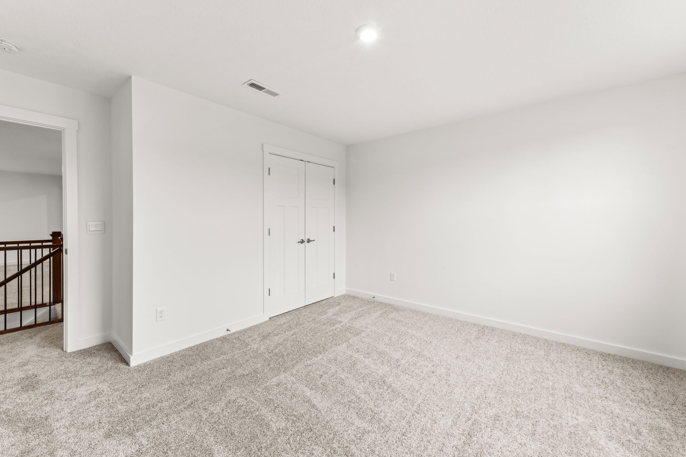 Empty white-walled room with plush carpet, recessed lighting, and a closed double-door closet adjacent to an open doorway with wooden railing.