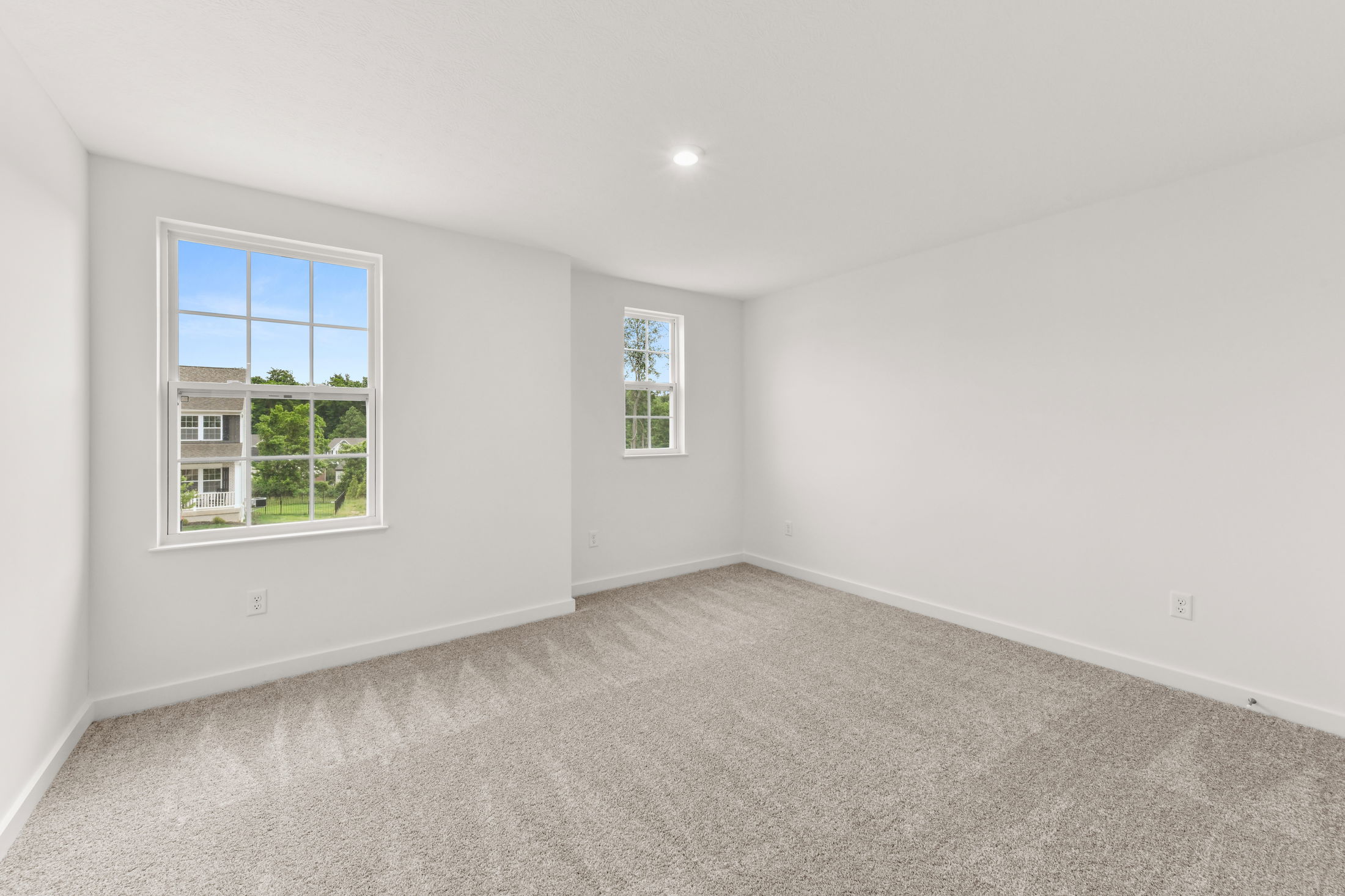 A bright, empty room with white walls, carpeted floors, and two windows showcasing a suburban view.