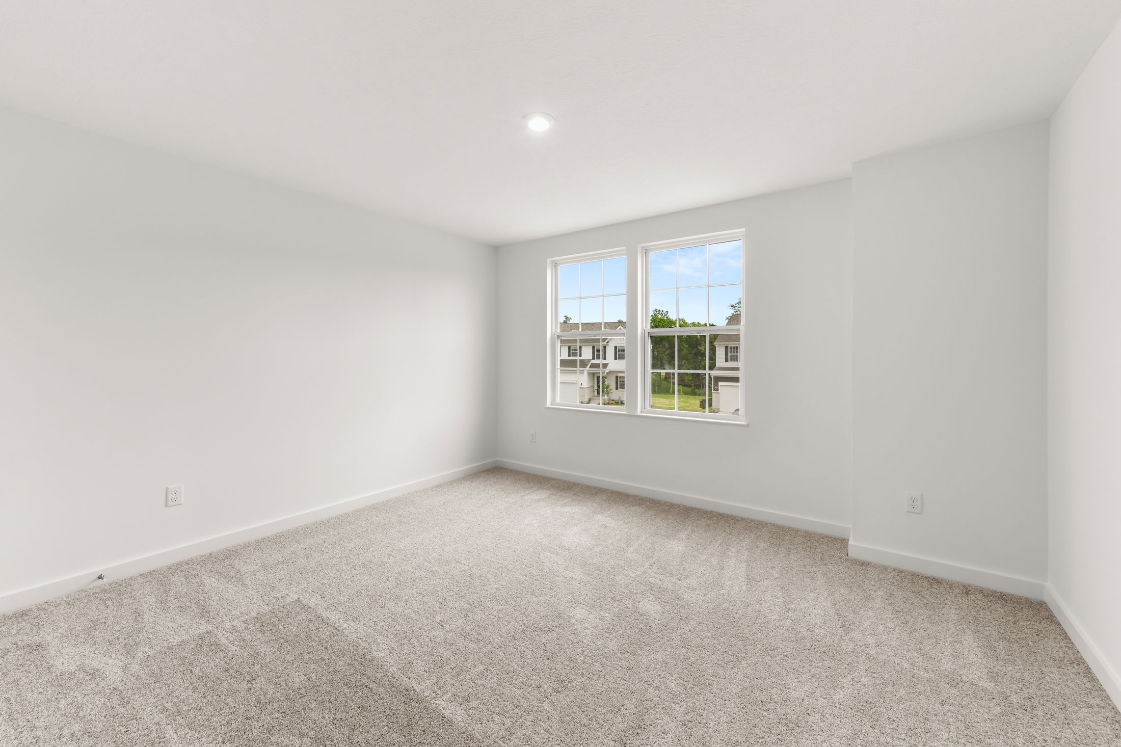 Bright, empty room with white walls, plush beige carpet, and a large window overlooking a residential neighborhood.