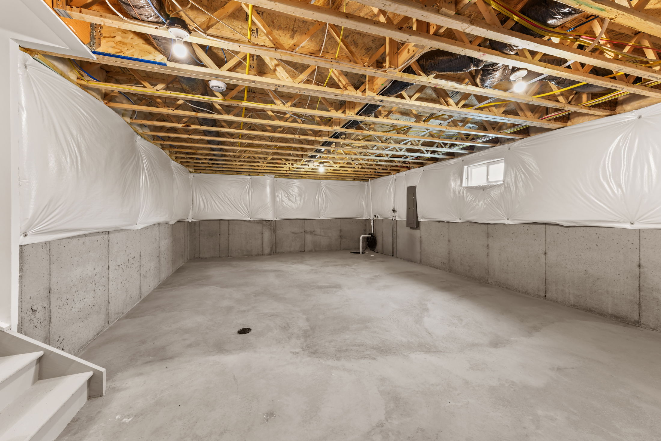 Spacious unfinished basement with exposed wooden ceiling beams, concrete walls, and plastic wall lining.