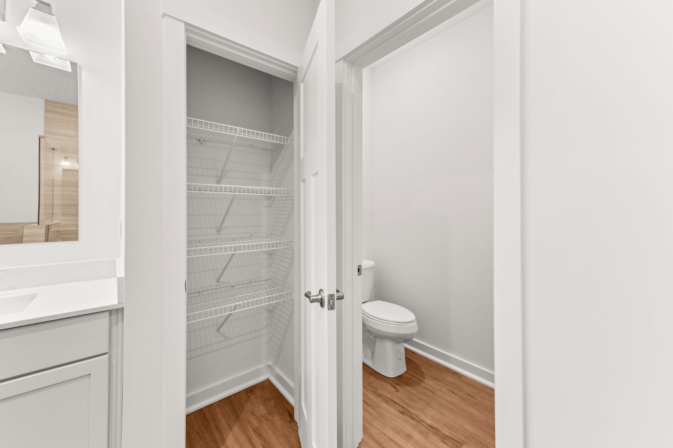 Modern bathroom with hardwood flooring showcasing a closet with wire shelving and a toilet in a white interior.