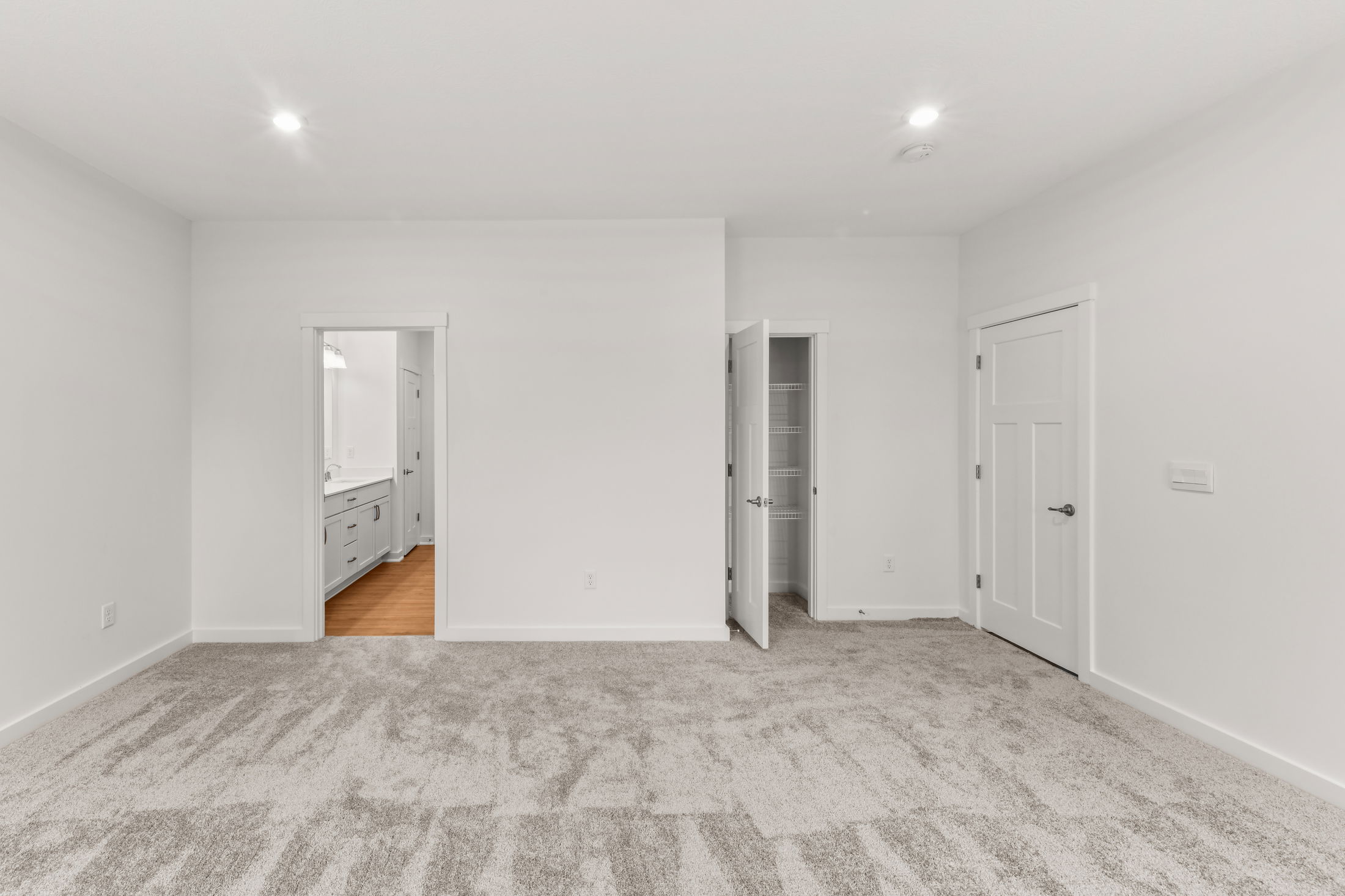 Empty, white-walled room with gray carpet, featuring two open doors leading to a bathroom and a closet.