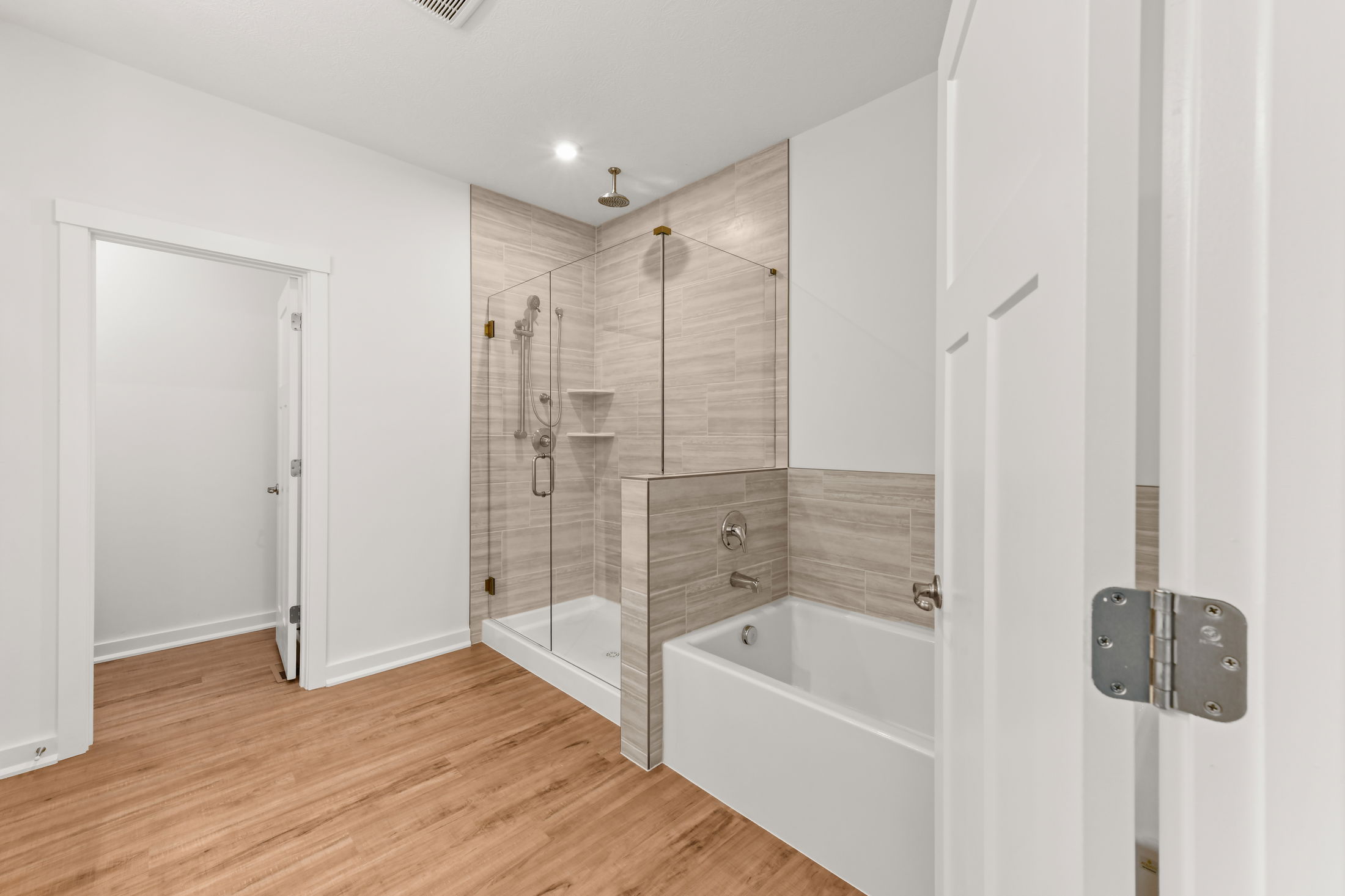 Modern bathroom with a glass-enclosed shower and adjacent bathtub, featuring wood-style flooring and beige wall tiles.