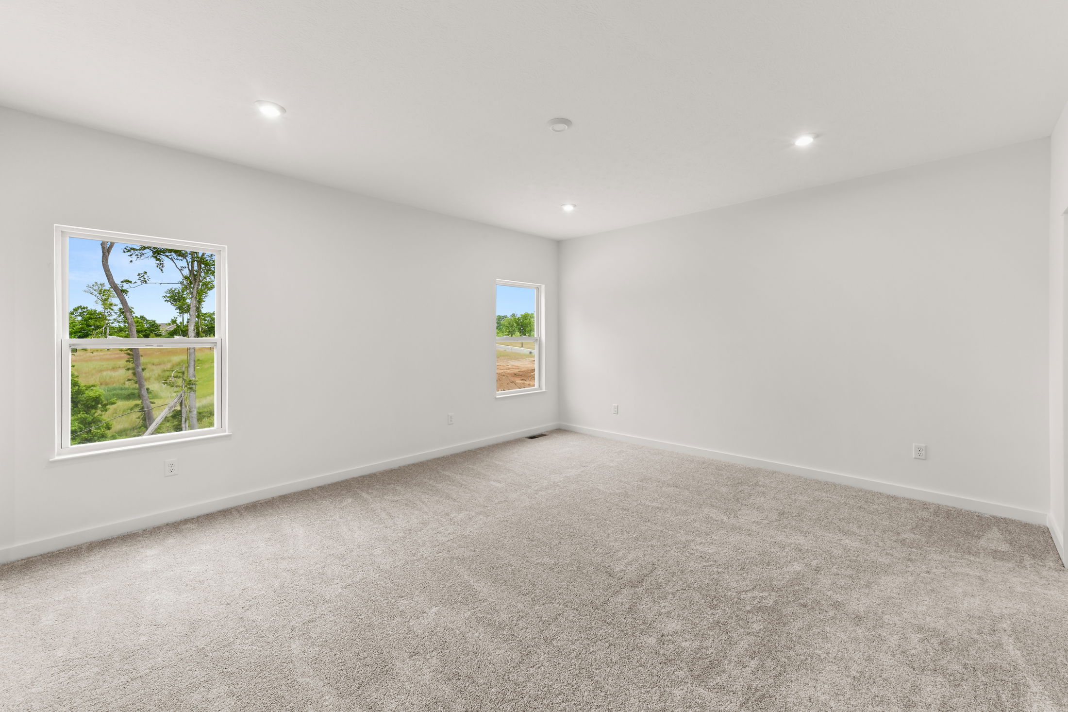 Spacious empty room with beige carpet, white walls, recessed lighting, and two windows offering a countryside view.