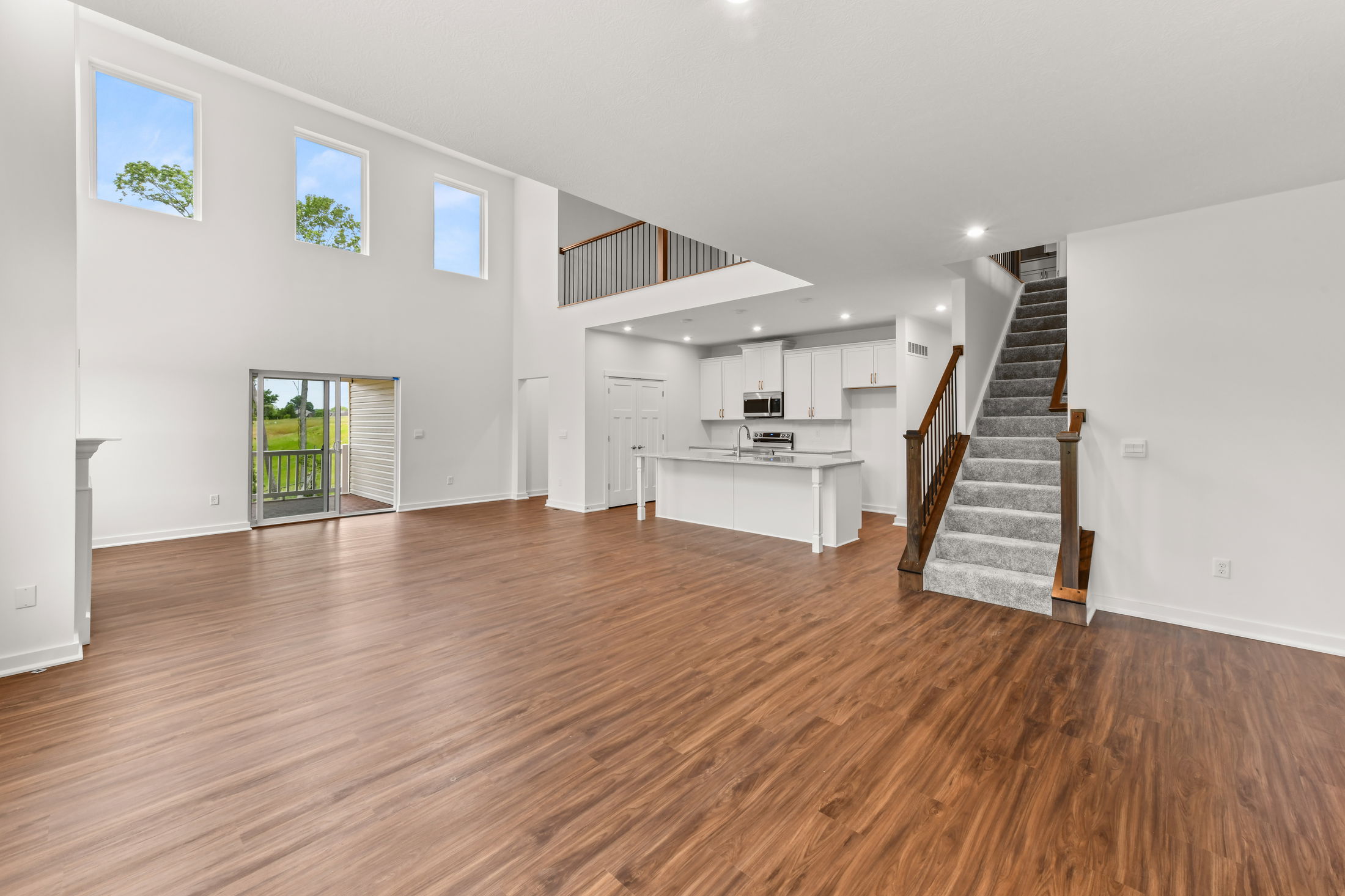 Spacious modern living room with high ceilings, hardwood floors, large windows, and an open-concept kitchen with white cabinetry and a staircase.
