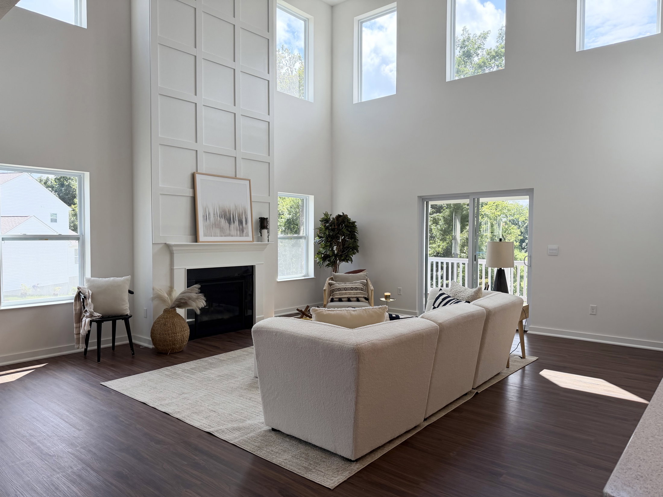 Bright living room featuring a white sectional sofa, a fireplace, and large windows with a view of greenery.