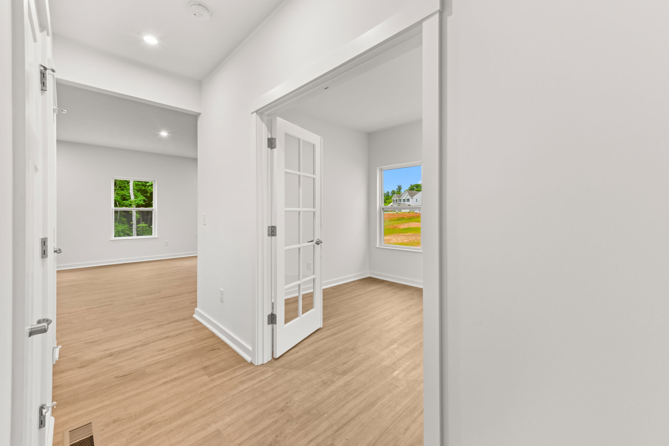 Bright, modern interior with white walls, wooden floors, and a French door leading to a room with a view of greenery outside.