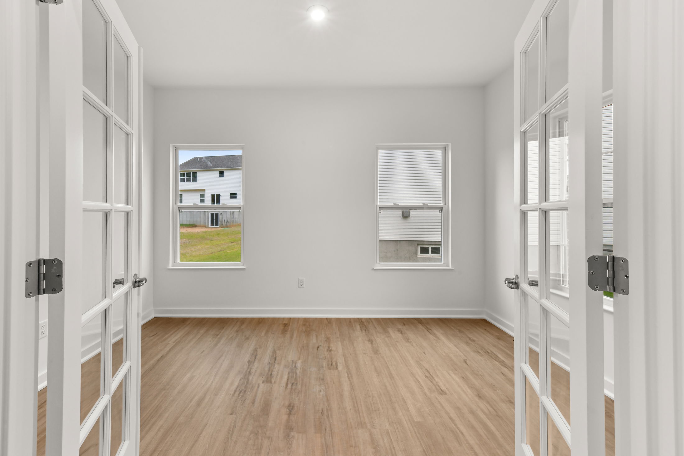 Bright, empty room with wooden floors and French doors, showcasing two windows with a view of neighboring houses.