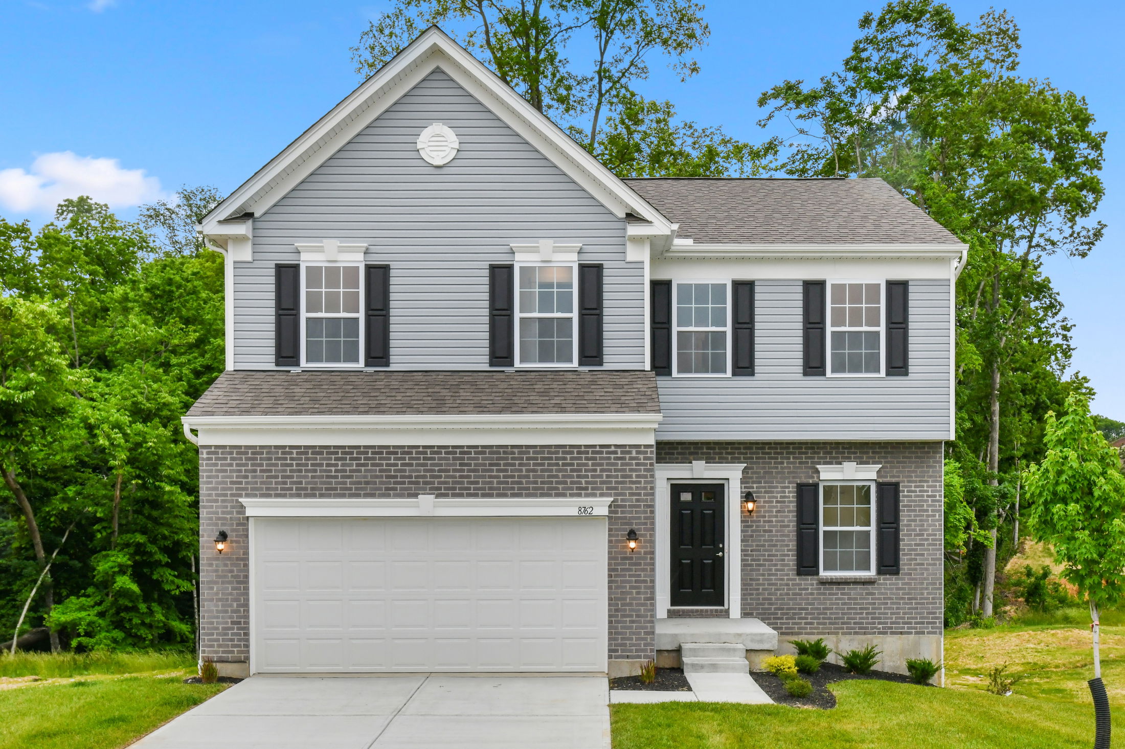 Contemporary two-story gray house with brick facade, black shutters, and a white garage door, set against a lush green backdrop.