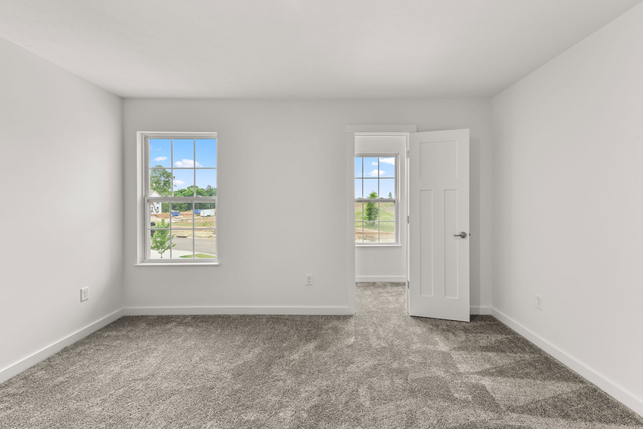 Empty room with gray carpet, white walls, two large windows, and an open white door showcasing natural light.