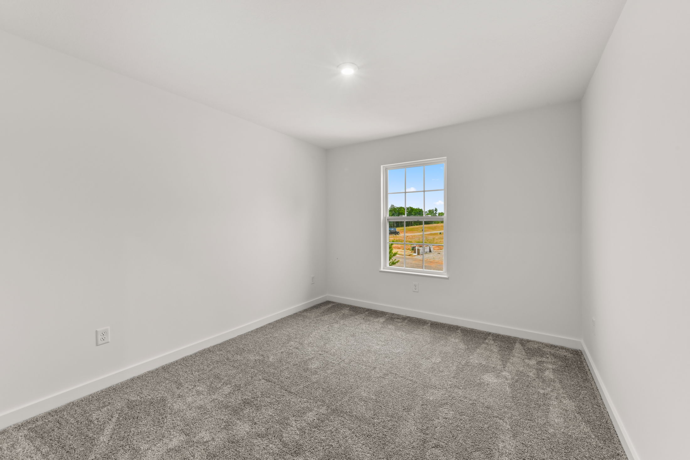 Empty room with gray carpet, white walls, and a bright window overlooking an outdoor landscape.