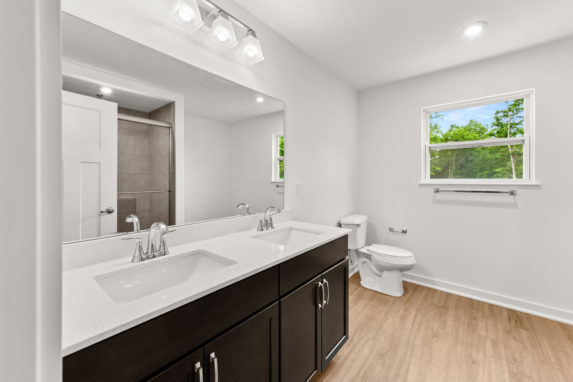 Modern bathroom with dual sinks, dark wood cabinets, white countertops, a toilet, and a large mirror reflecting a shower and light fixture.
