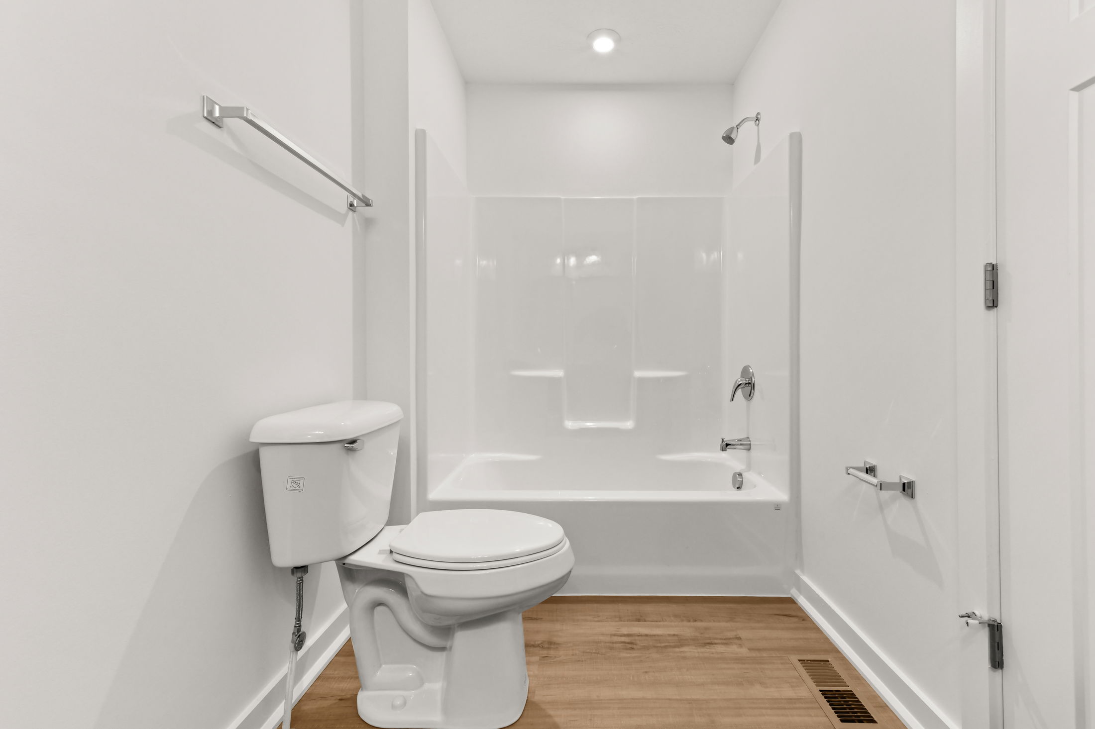 Modern minimalist bathroom with white fixtures, featuring a toilet and a shower-tub combo against a neutral wall with wood flooring.