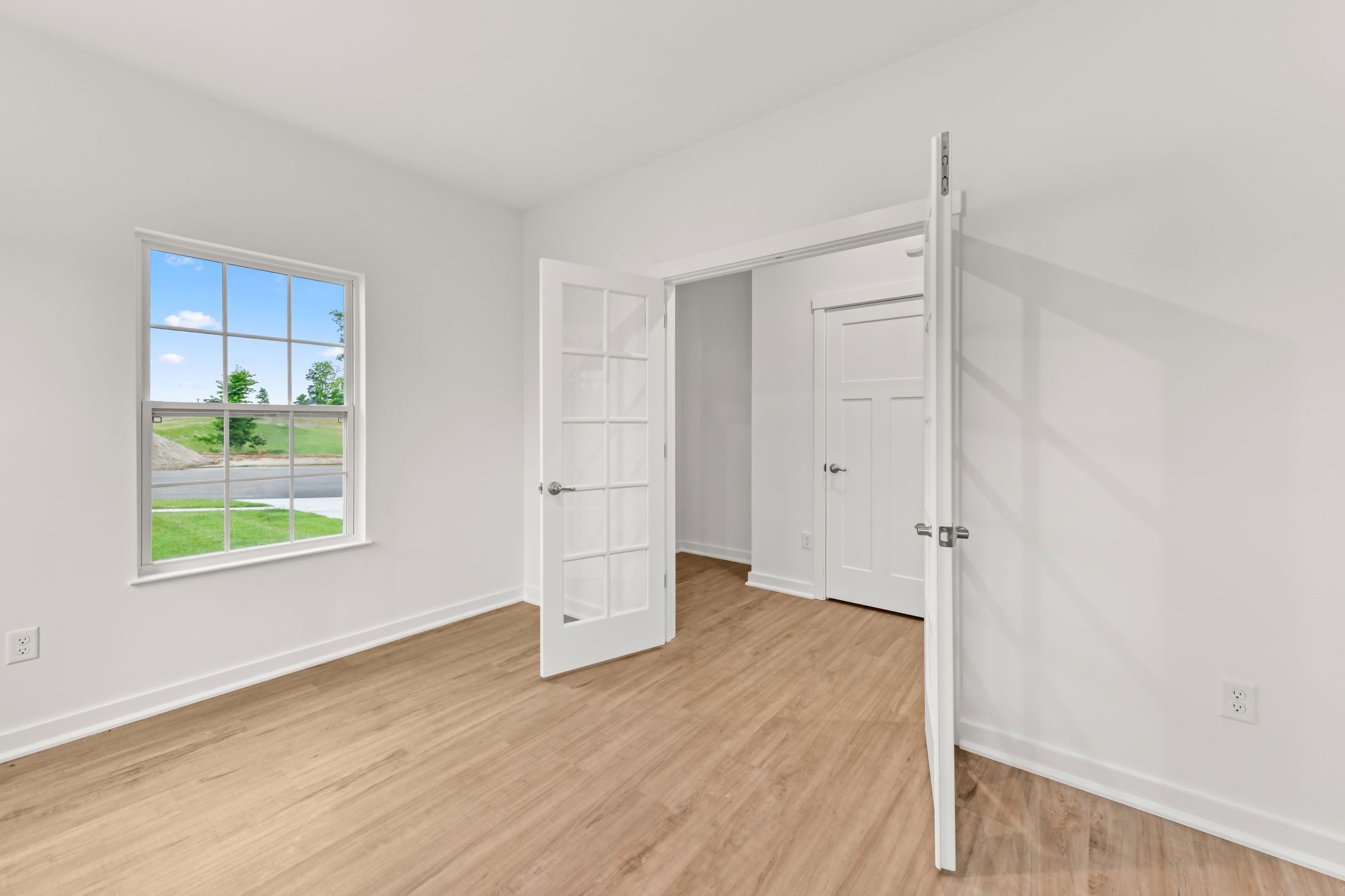 Bright, empty room with wooden floors, white walls, French doors, and a window overlooking a green lawn.