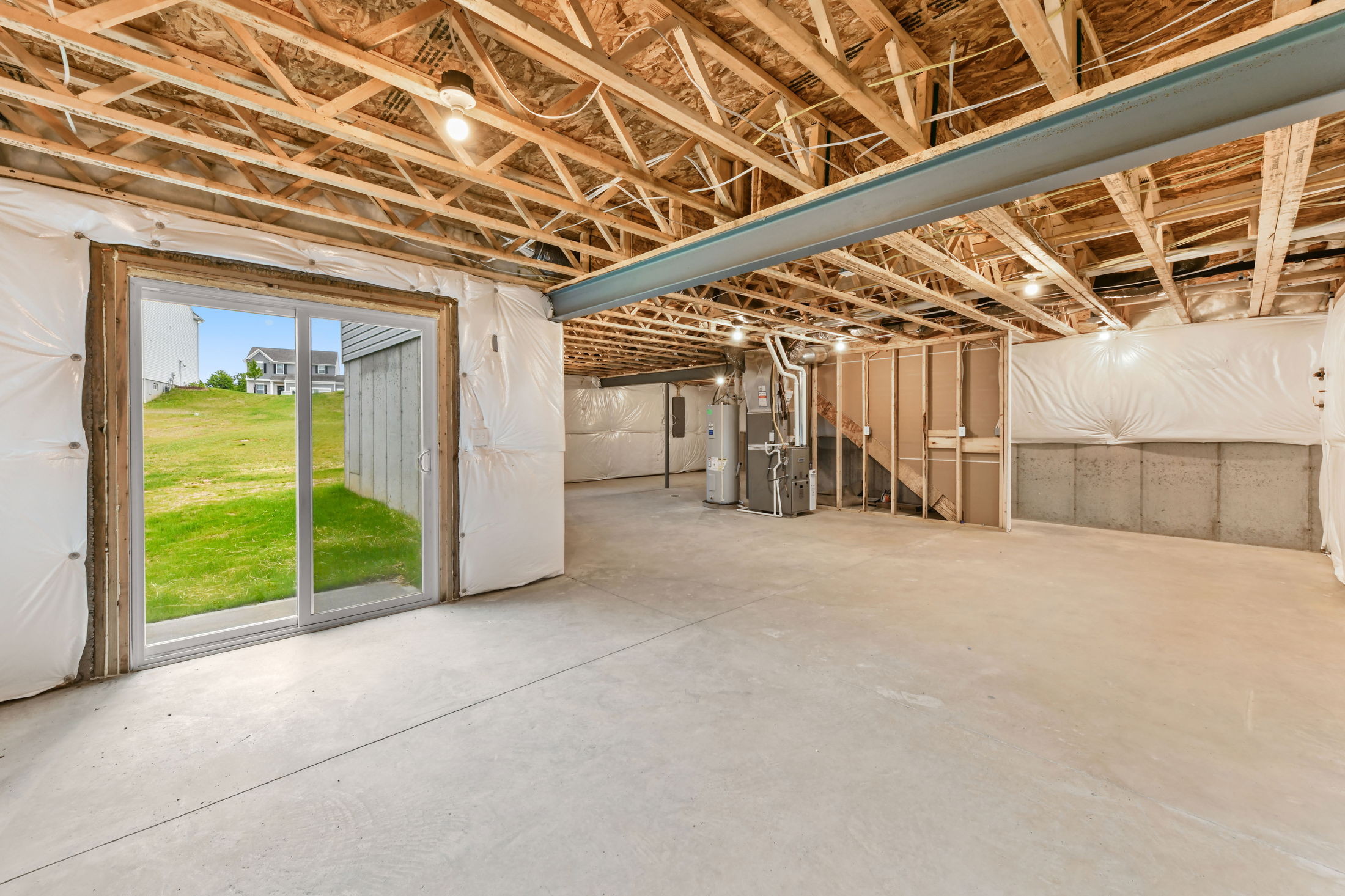 Unfinished basement with exposed wooden beams, concrete floors, and sliding glass door leading to a grassy backyard.