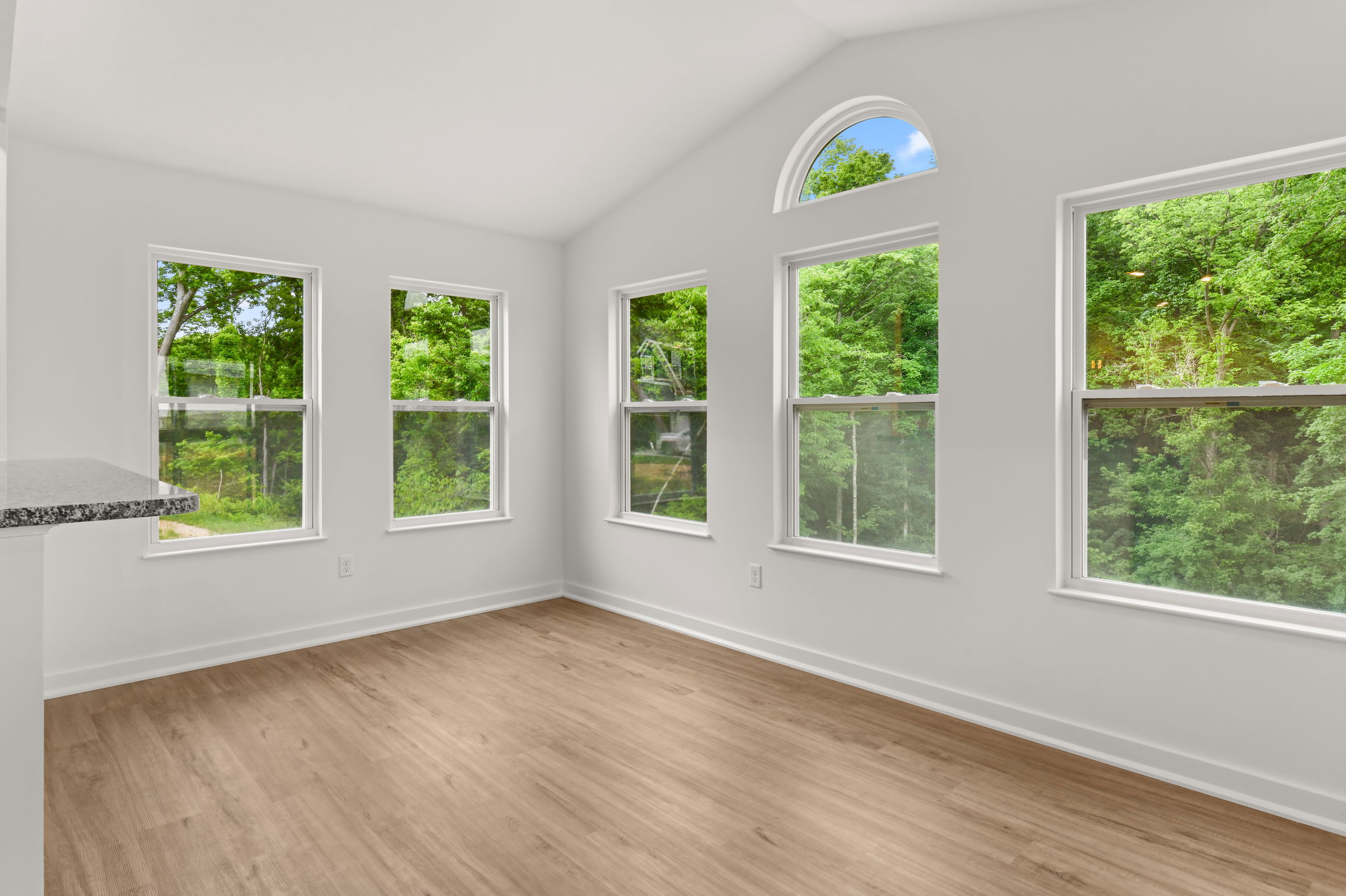 Bright, empty room with large windows showcasing a lush green forest view and wood flooring.
